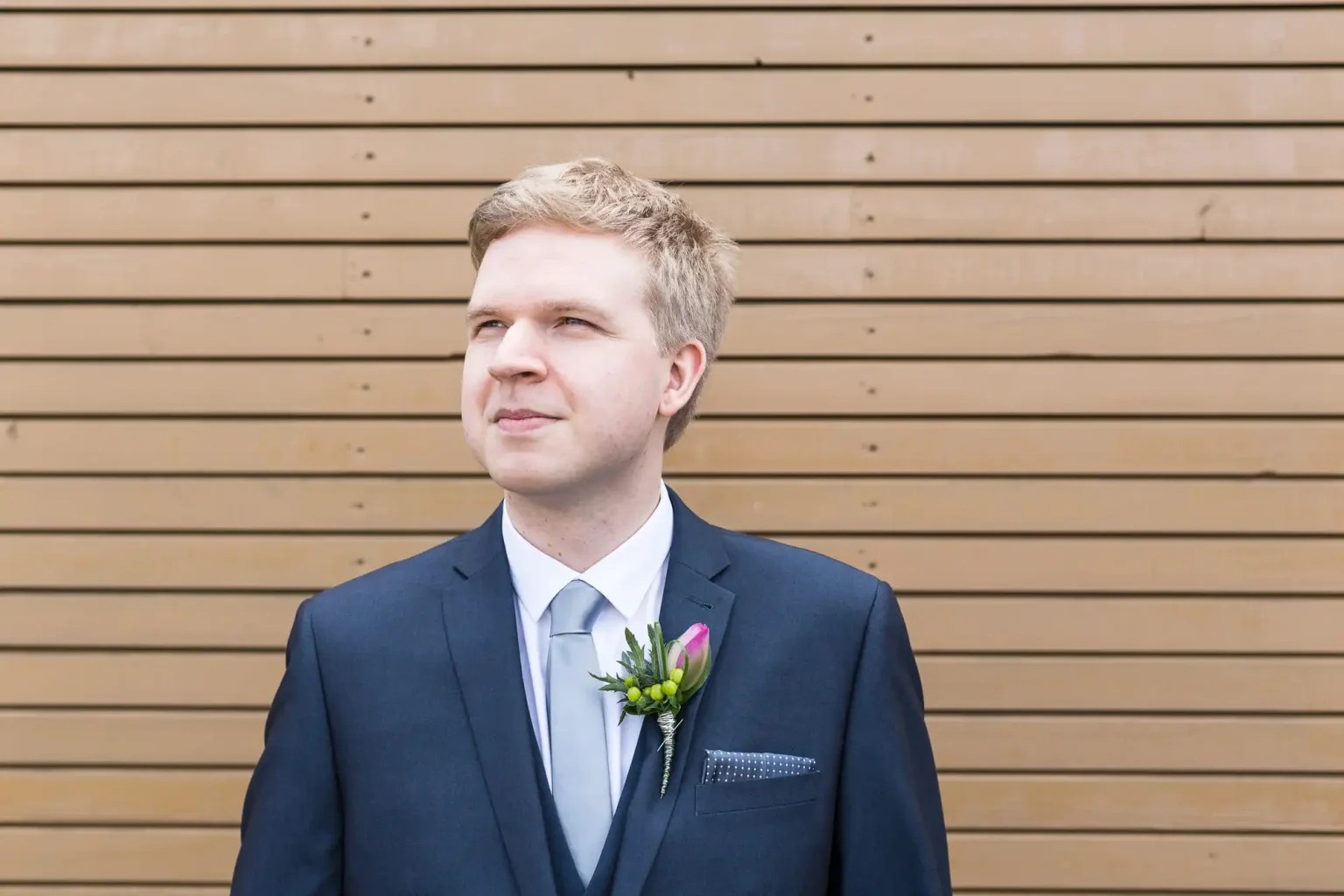A man in a dark suit with a boutonniere, looking to the side thoughtfully, standing in front of a wooden slat background.