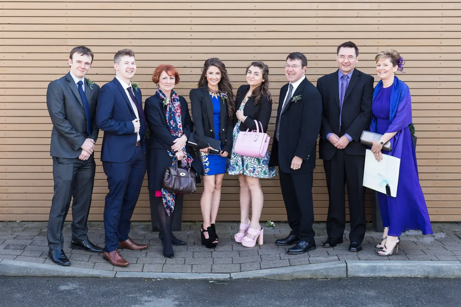Eight people dressed in formal attire standing in front of a wooden wall, smiling for a group photo.