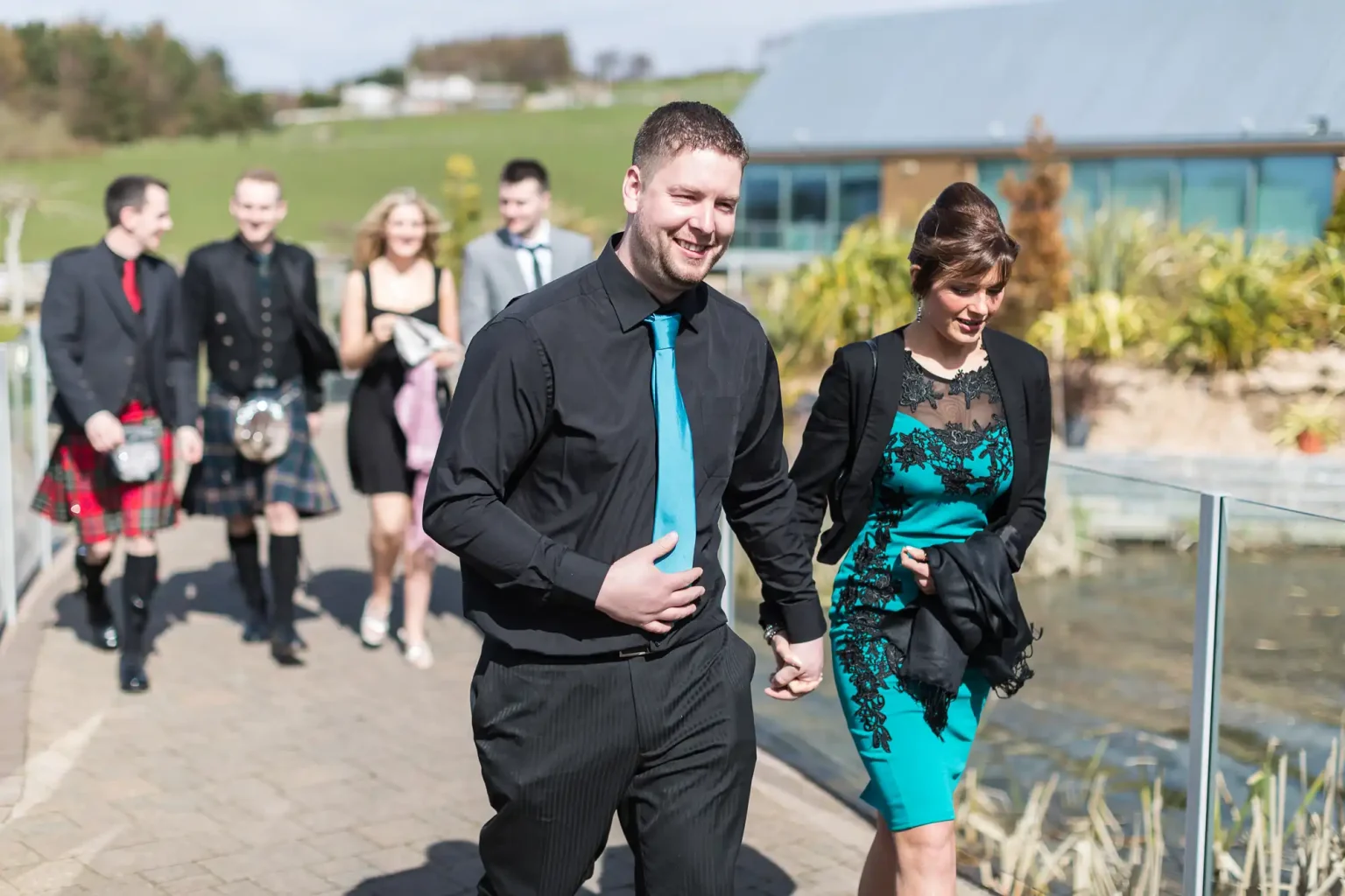 A man in a black suit and a woman in a teal dress walk together at an event, smiling, with other guests in various attire in the background.