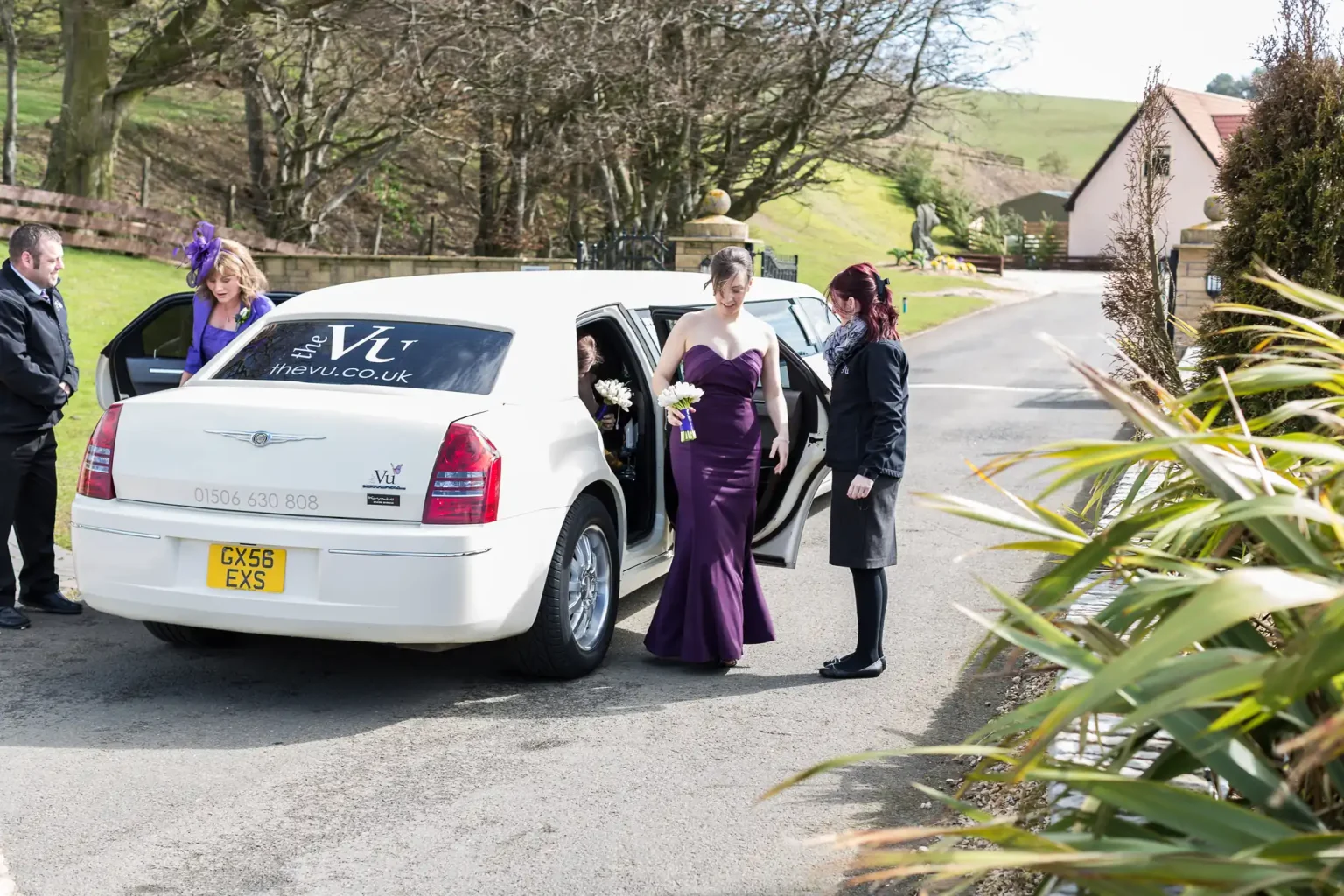 People and a chauffeur standing beside a white stretch limousine at a countryside event, with hills in the background.