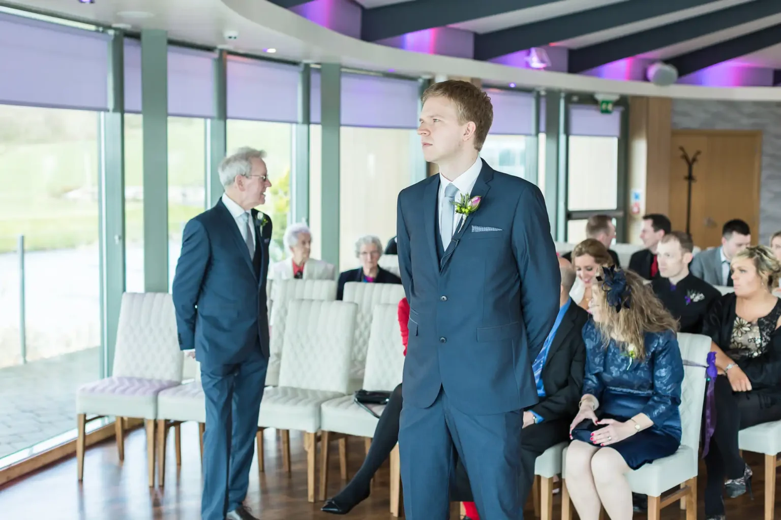 A groom standing in a ceremony hall with seated guests, conversing with an older man, with large windows showing a green landscape outside.