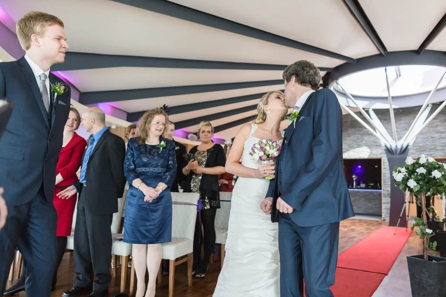 Newlyweds kissing at their wedding ceremony, surrounded by smiling guests in a festively decorated room with a vaulted ceiling.