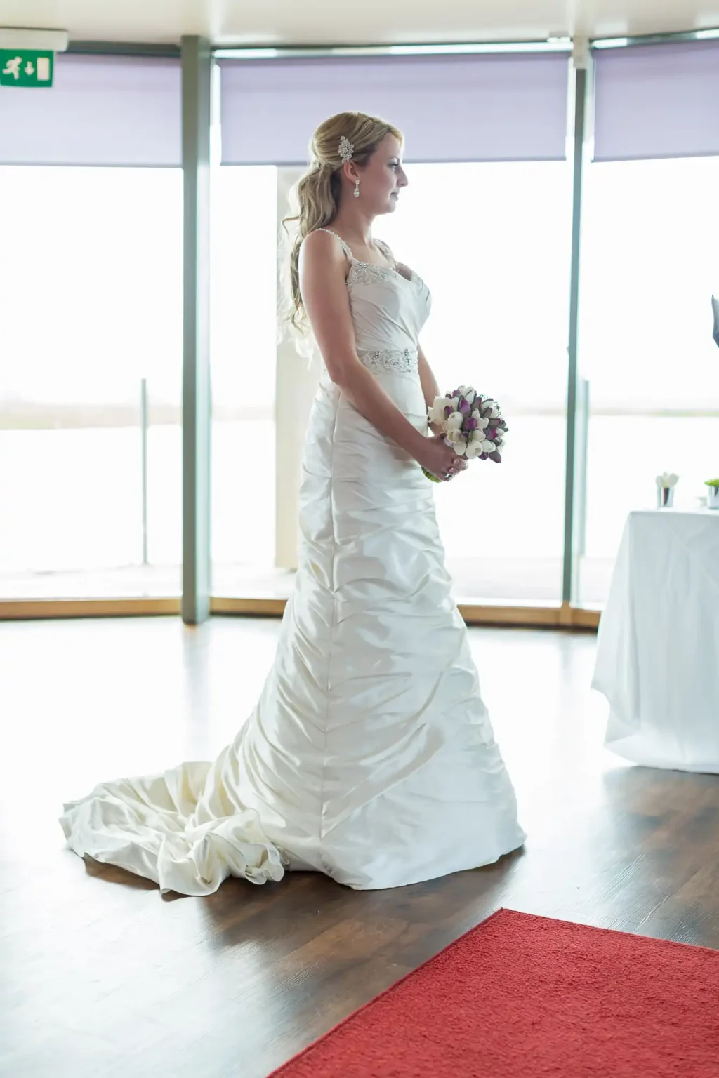 A bride in a white gown holding a bouquet, standing by large windows in a bright room.