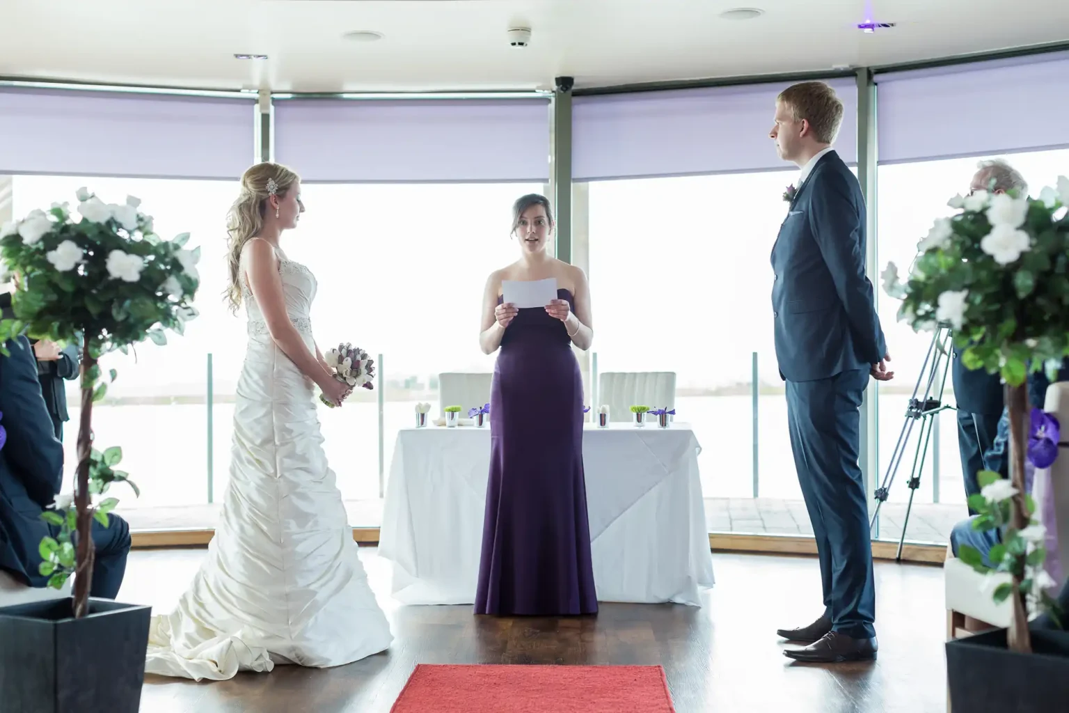 A bride and groom standing face to face at their wedding ceremony, with an officiant reading from a paper, inside a room decorated with white flowers.