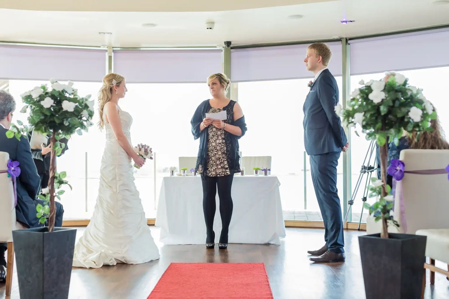 A wedding ceremony inside a room with large windows; a bride and groom facing each other, with an officiant reading from a note.