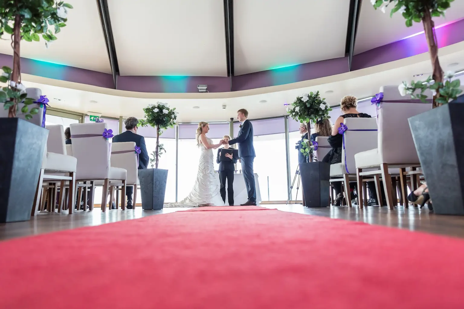 A bride and groom holding hands during a wedding ceremony in a modern room with guests seated around them and a red carpet aisle.