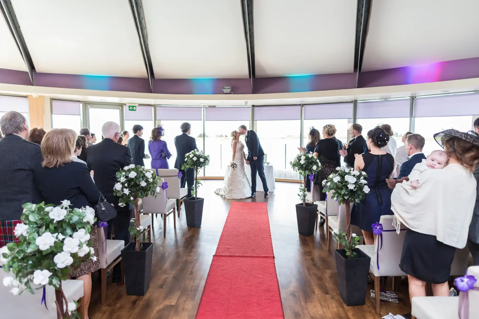 Wedding ceremony in Waterlily at The Vu, with guests facing a couple exchanging vows at the end of a red carpet, surrounded by floral decorations.