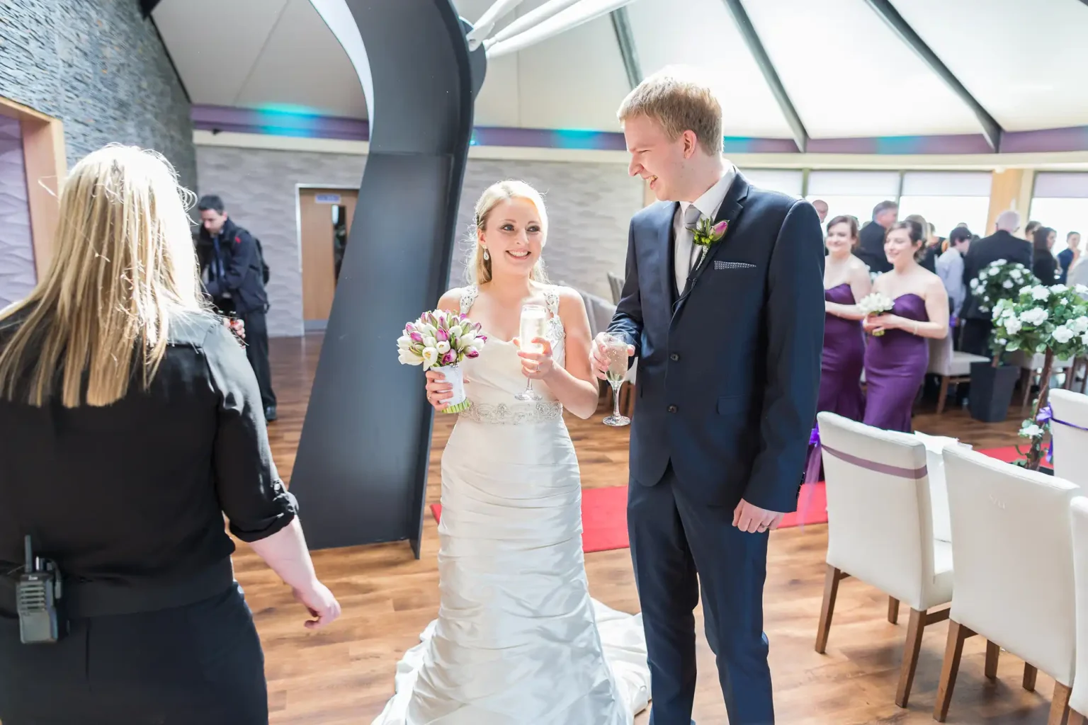A bride and groom walk hand in hand, smiling as they enter a wedding reception. guests in formal attire are visible in the background.