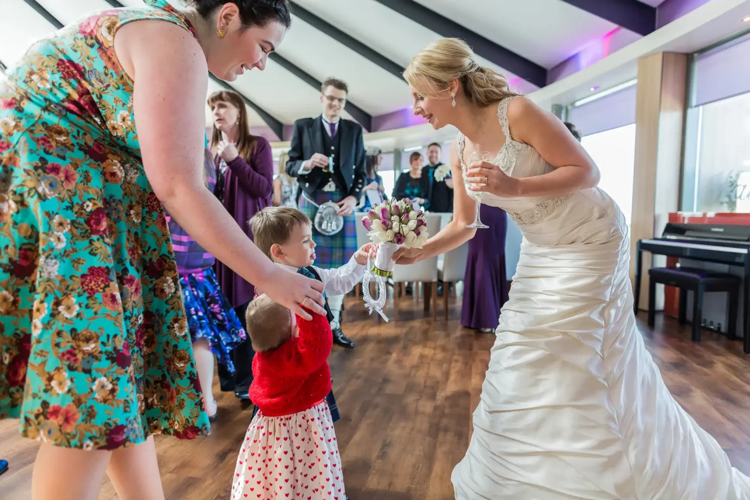 A bride in a white dress gives a ribbon to a small child at a wedding, with guests looking on in an elegantly decorated room.
