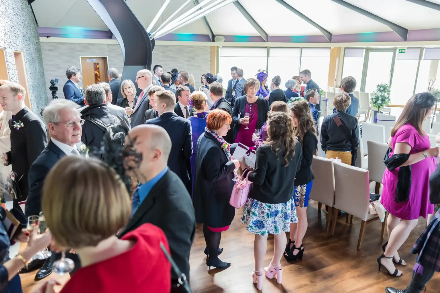 A group of elegantly dressed guests mingling at an indoor event, with natural light streaming in from large windows.