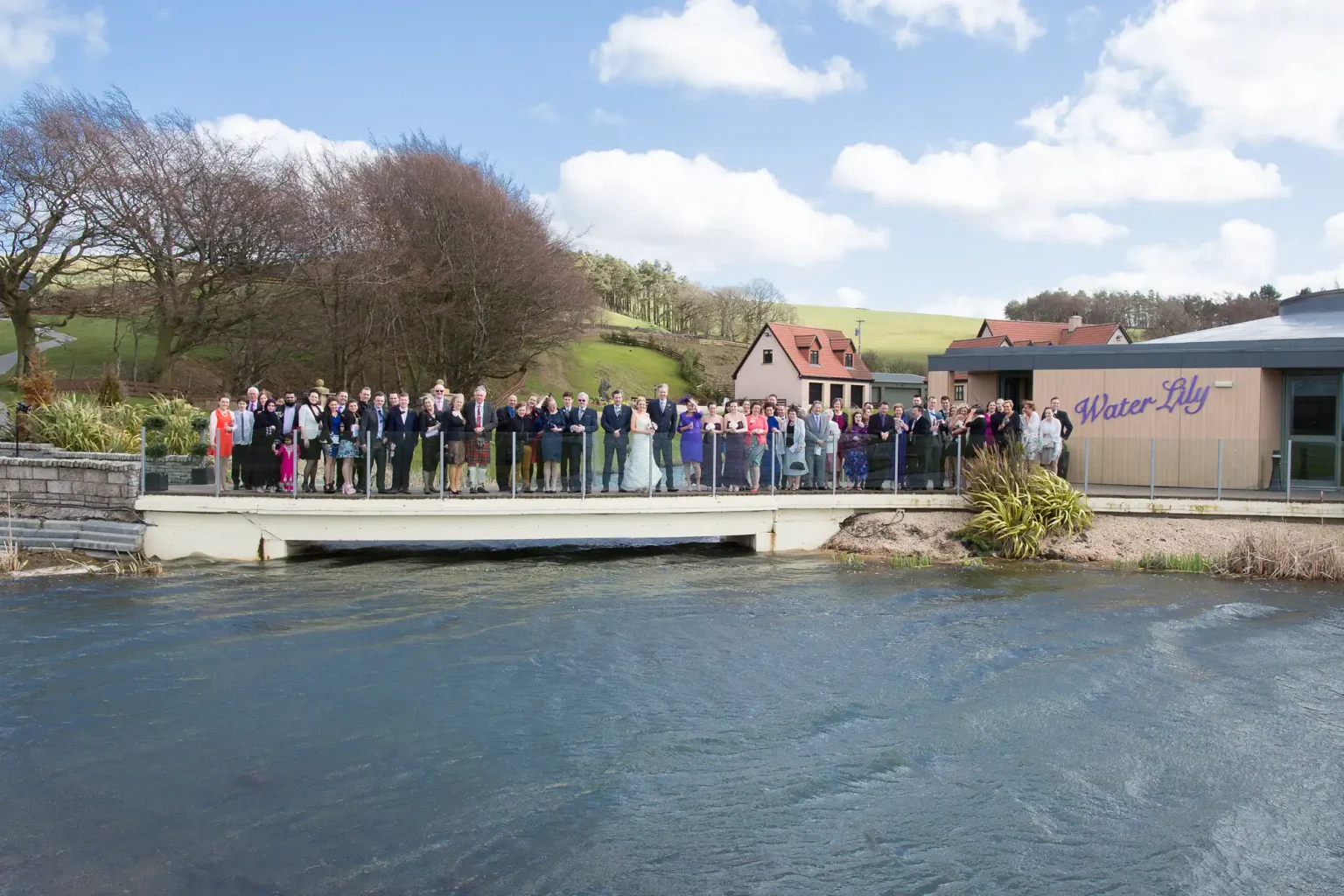 A group of people gathered on a deck by a river outside the water-lily building on a sunny day.