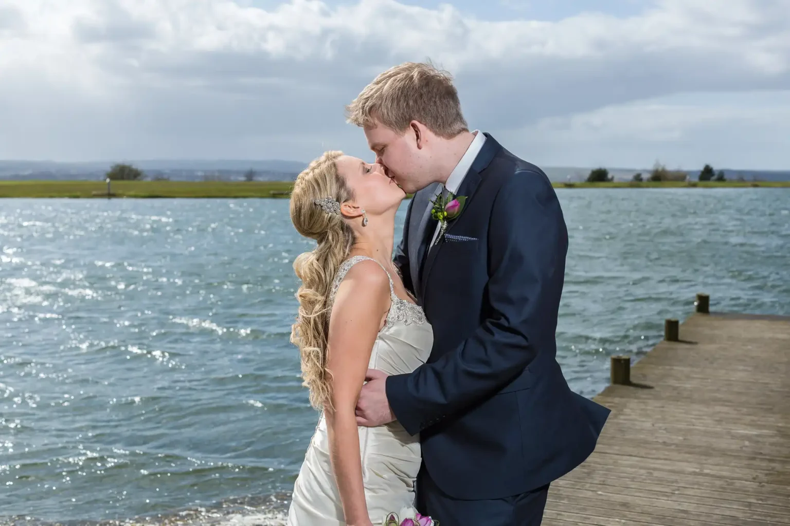 A bride and groom kissing on a wooden dock by a lake under a cloudy sky.