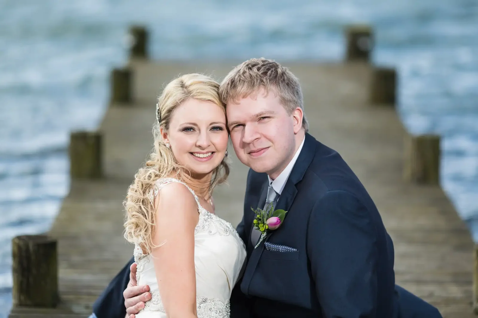 A bride and groom smiling, seated closely together on a wooden pier overlooking water, with cloudy skies above.
