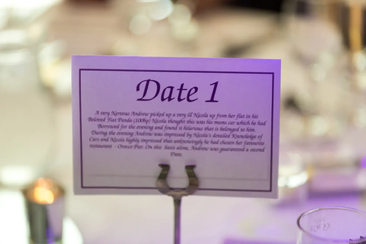 A purple table card labeled "date 1" with details of a memorable date printed on it, stands on a metallic holder, set against a blurred background at an event.