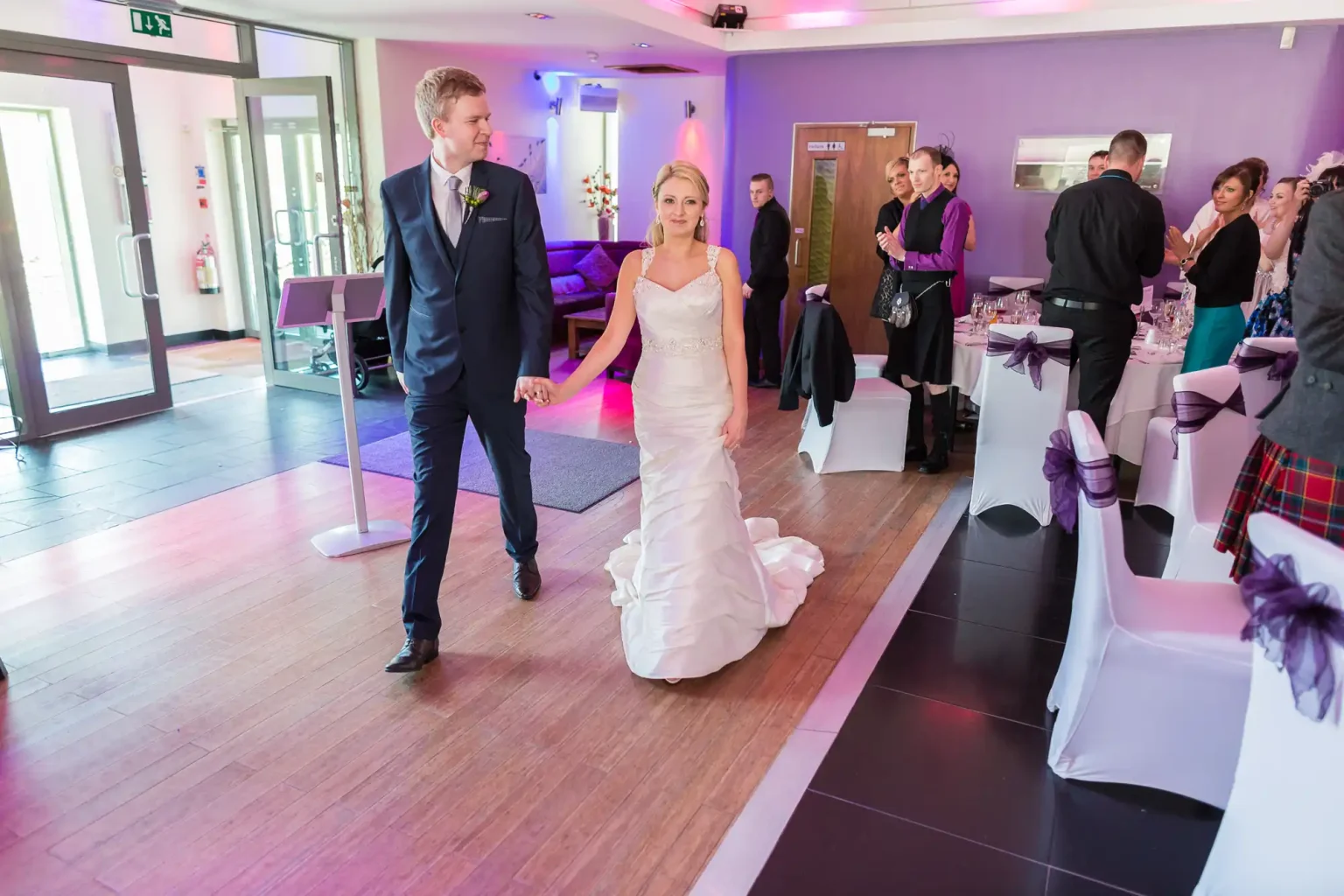 A newlywed couple walks hand in hand through a wedding venue, smiling as guests look on. the bride is in a white gown and the groom in a suit.