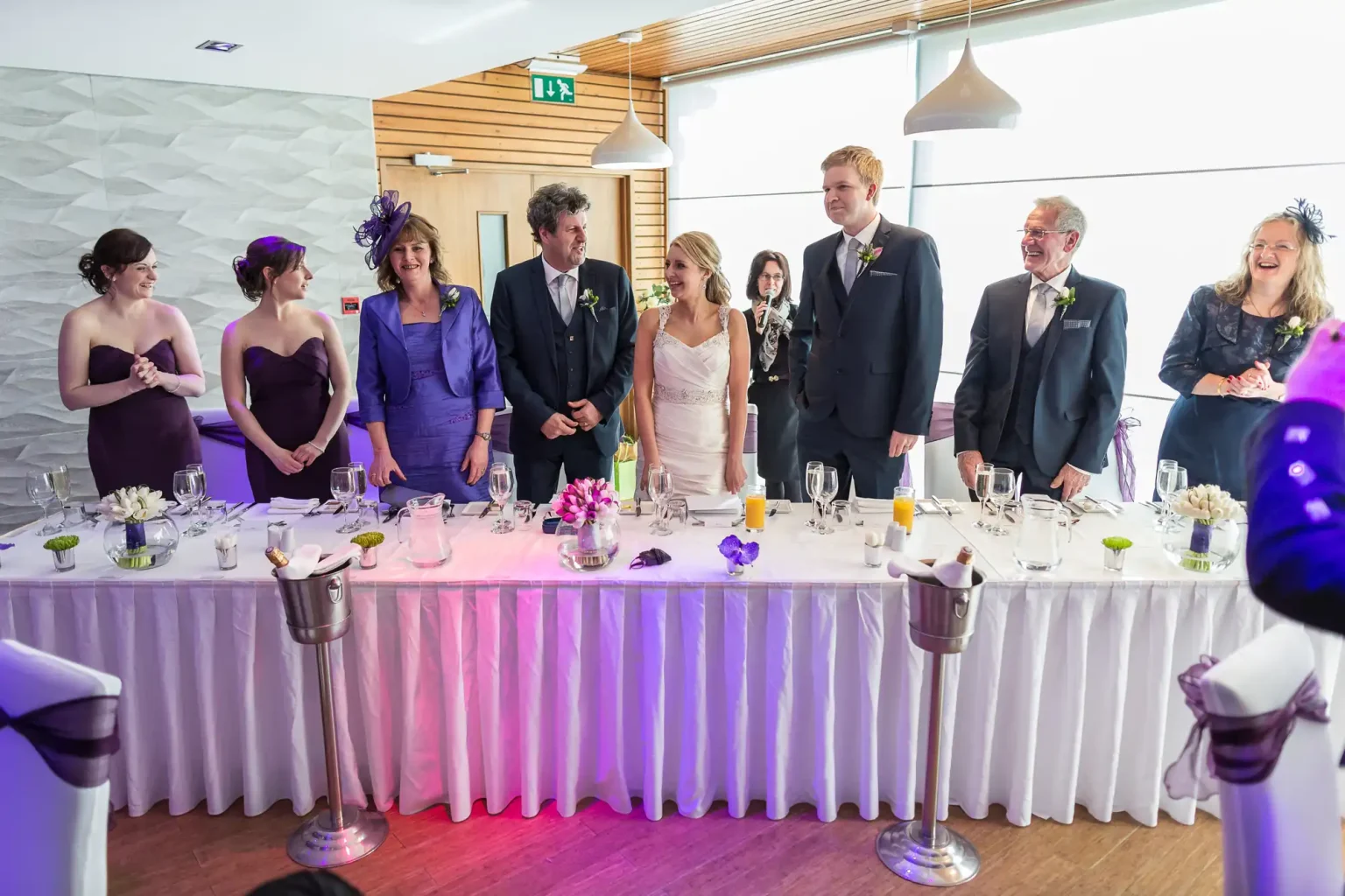 A wedding reception with a bride and groom standing at a decorated table, surrounded by guests in formal attire, smiles all around.