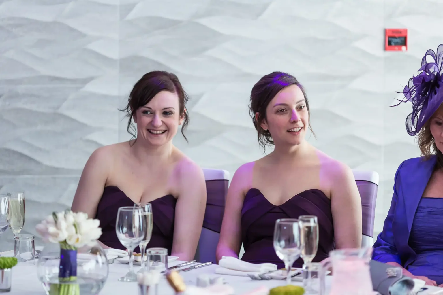 Two women in purple dresses smiling at a formal table setting with wine glasses and a white floral centerpiece, seated next to a woman in a blue hat.