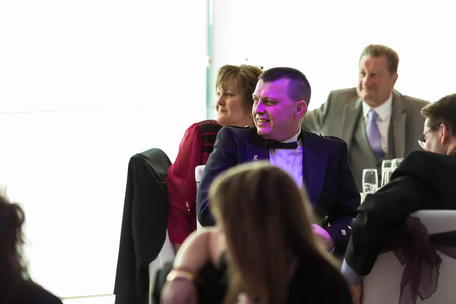 A man in a dark suit laughs among seated guests at a formal event, with a woman in a red jacket beside him, and another man smiling in the background.