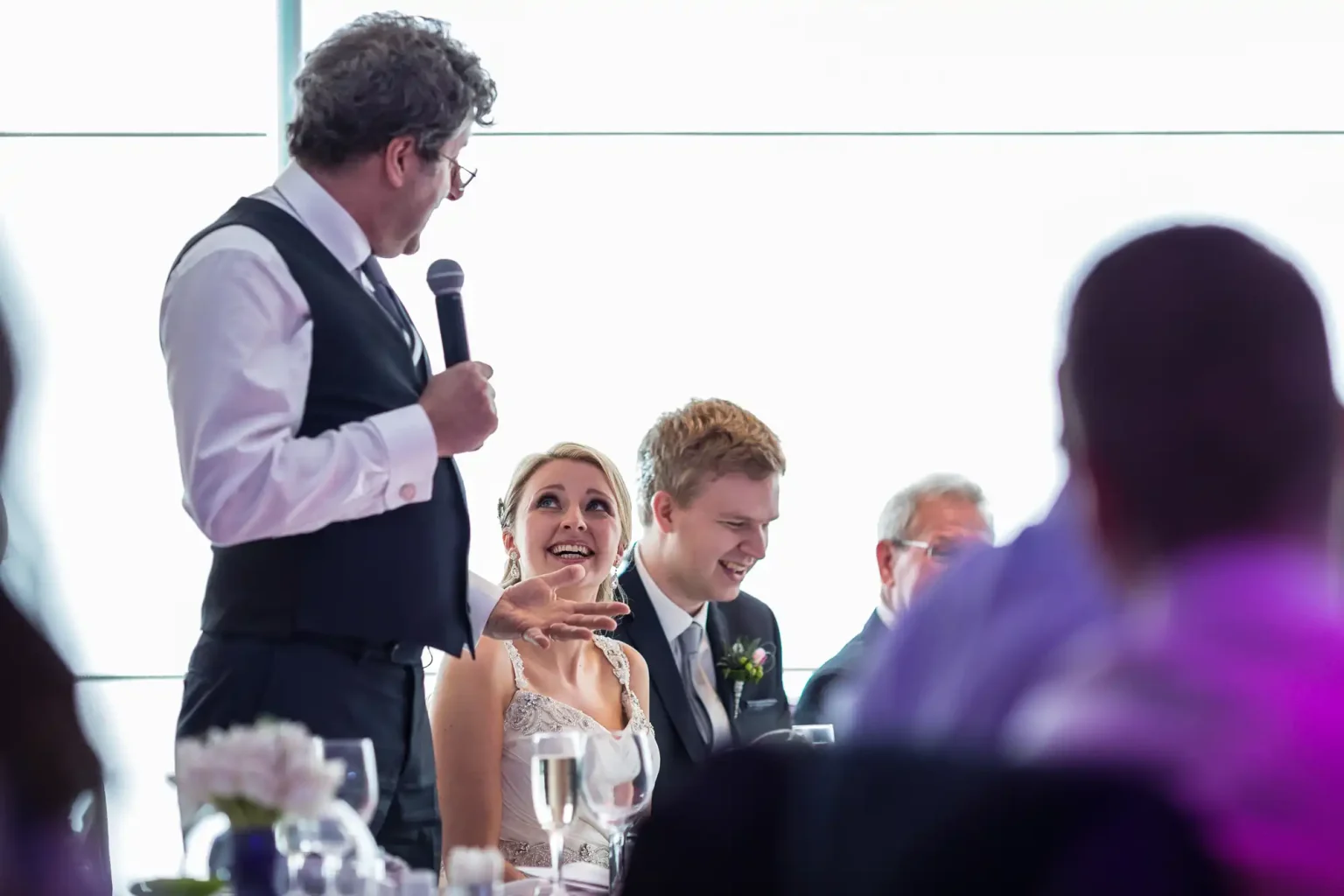A bride and groom laugh joyously at a wedding reception as an older man, holding a microphone, speaks to them and the guests.