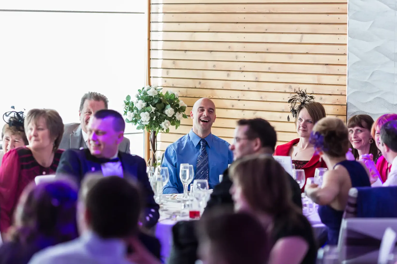 A man laughing joyfully at a table during a vibrant indoor gathering with several guests engaged in conversations around him.