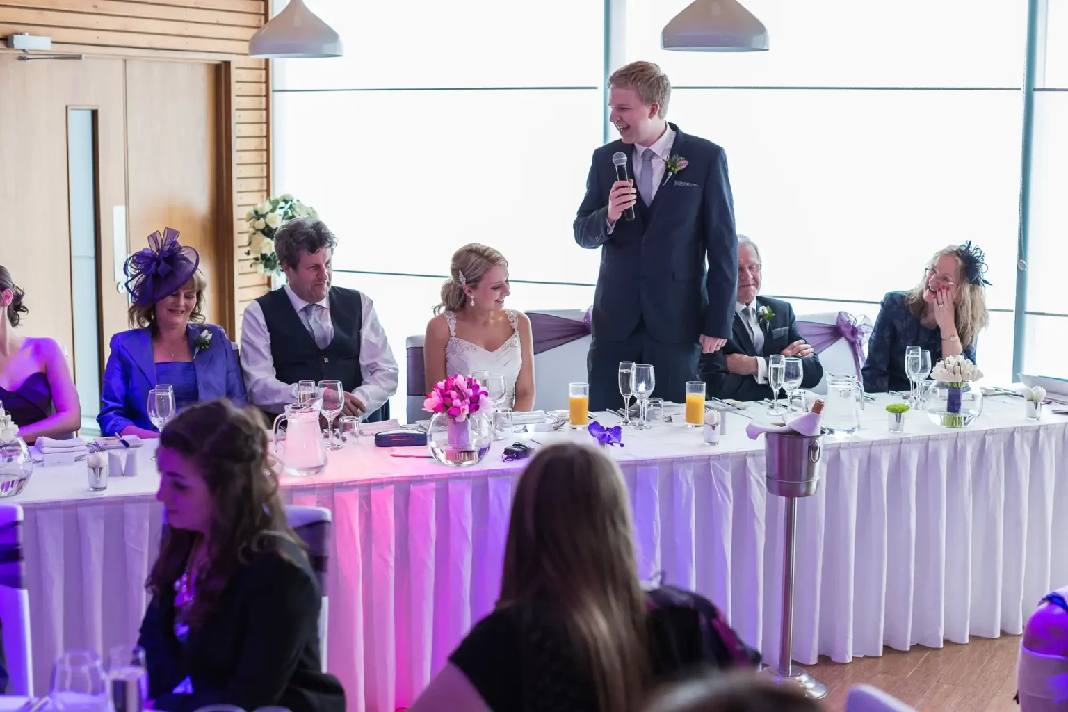 A young man gives a speech at a wedding reception, addressing attendees seated at decorated tables in an elegantly lit room.