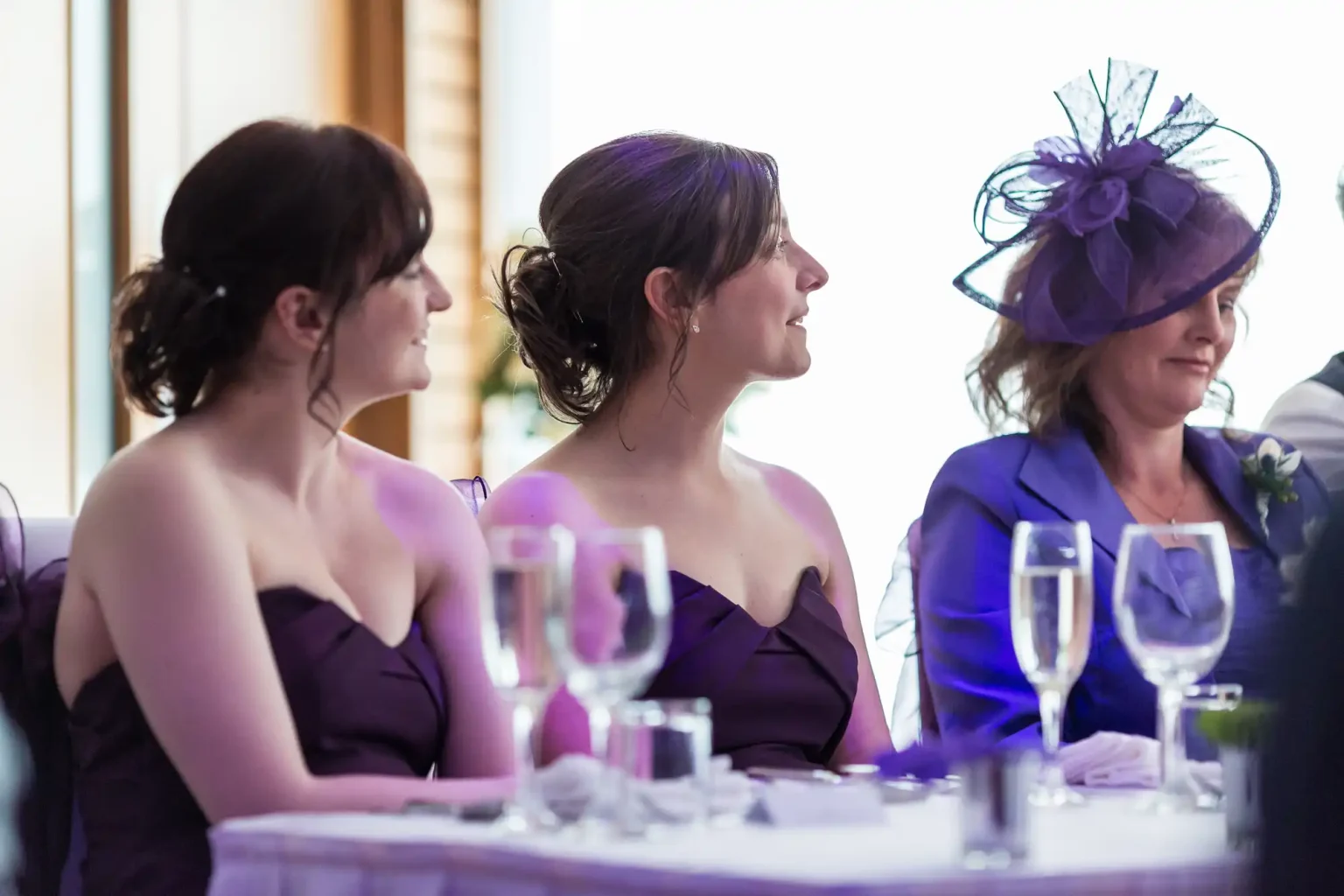 Three women in formal attire, two in purple dresses and one in a purple hat, sitting at a dining table during an event.