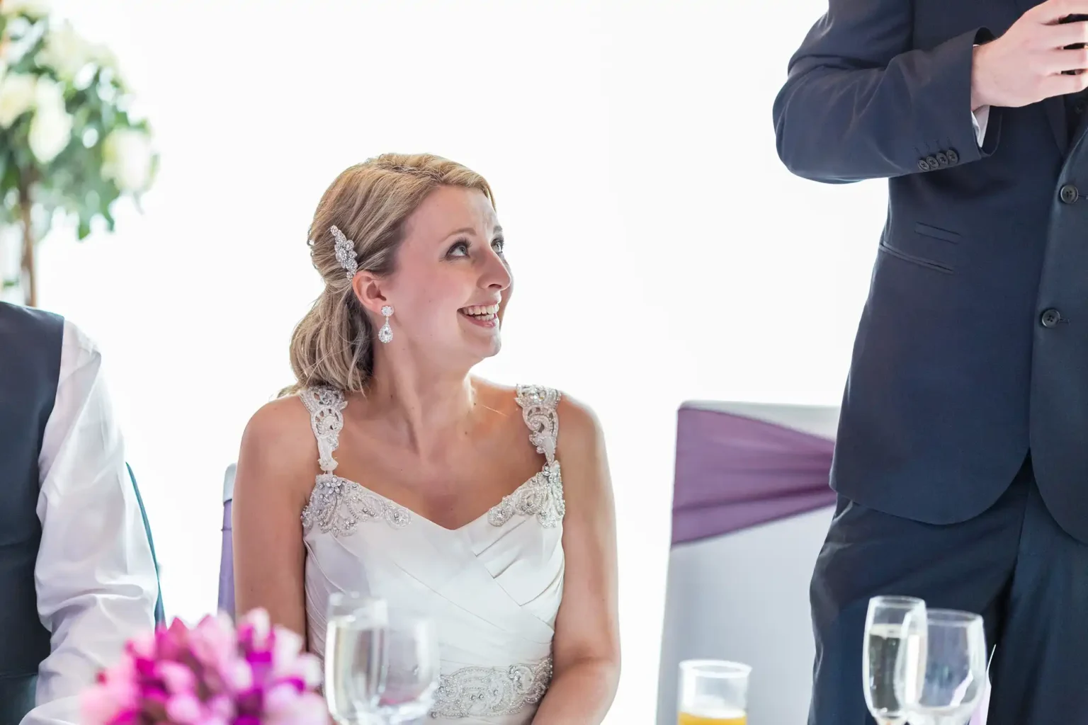 A bride in a lace wedding gown looks up smiling, seated at a table during a wedding reception, with a man standing beside her partially shown.