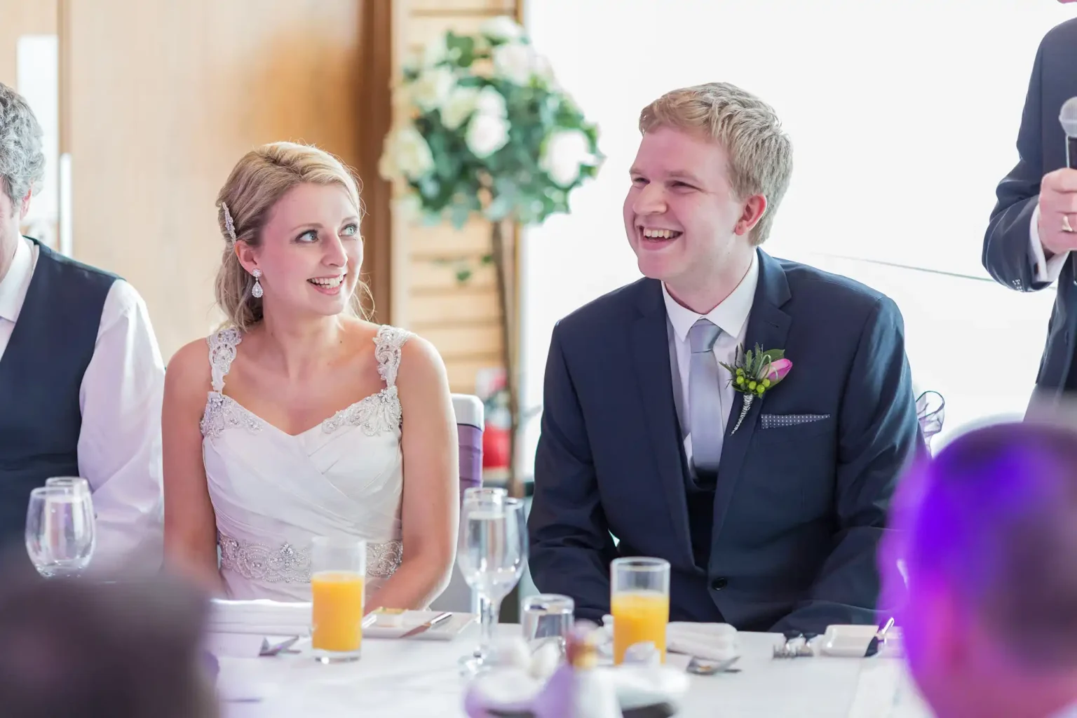 Bride and groom smiling and sitting at a wedding reception table, surrounded by guests. bright, well-lit room.