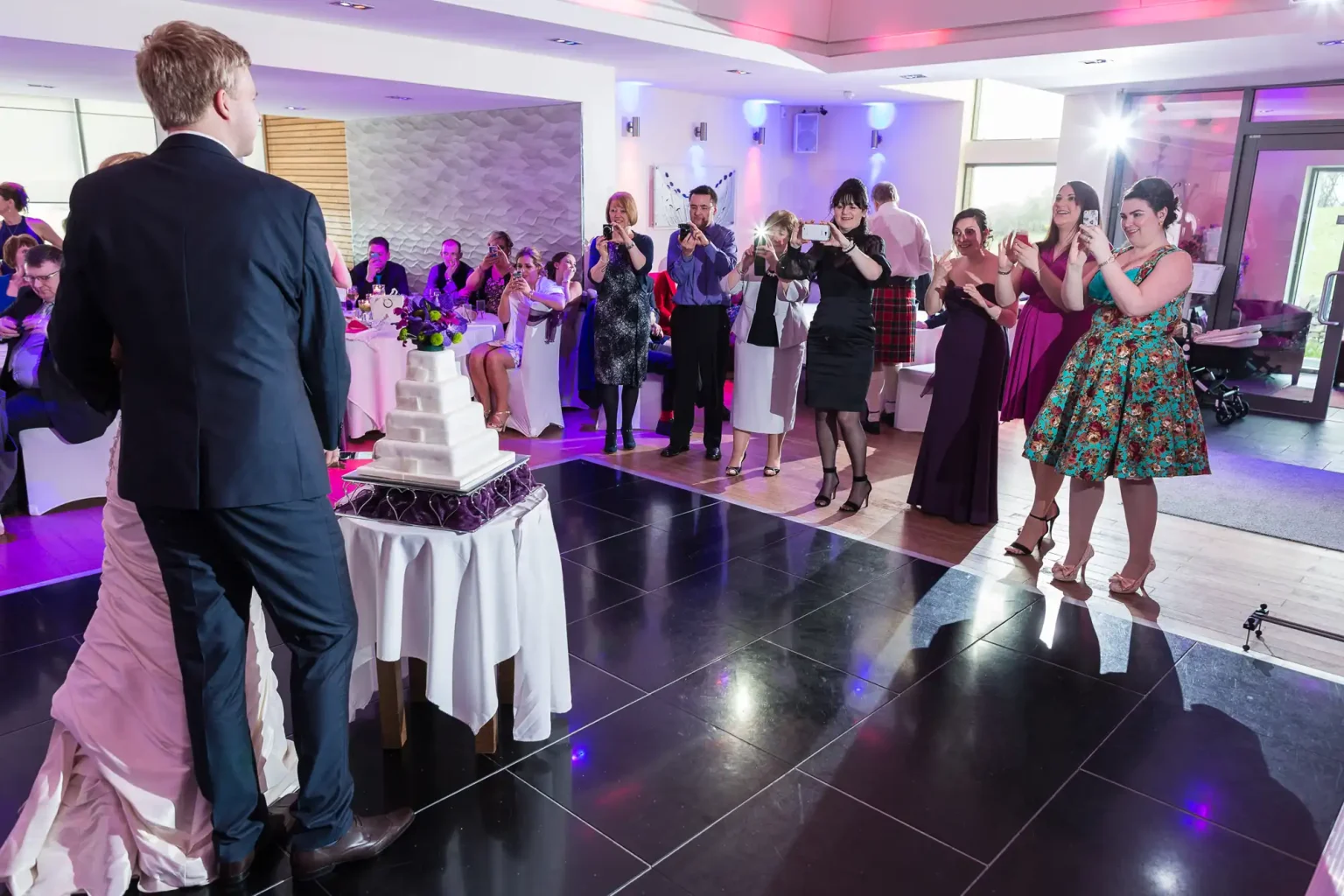 Wedding guests applauding a bride and groom near a cake in a banquet hall.