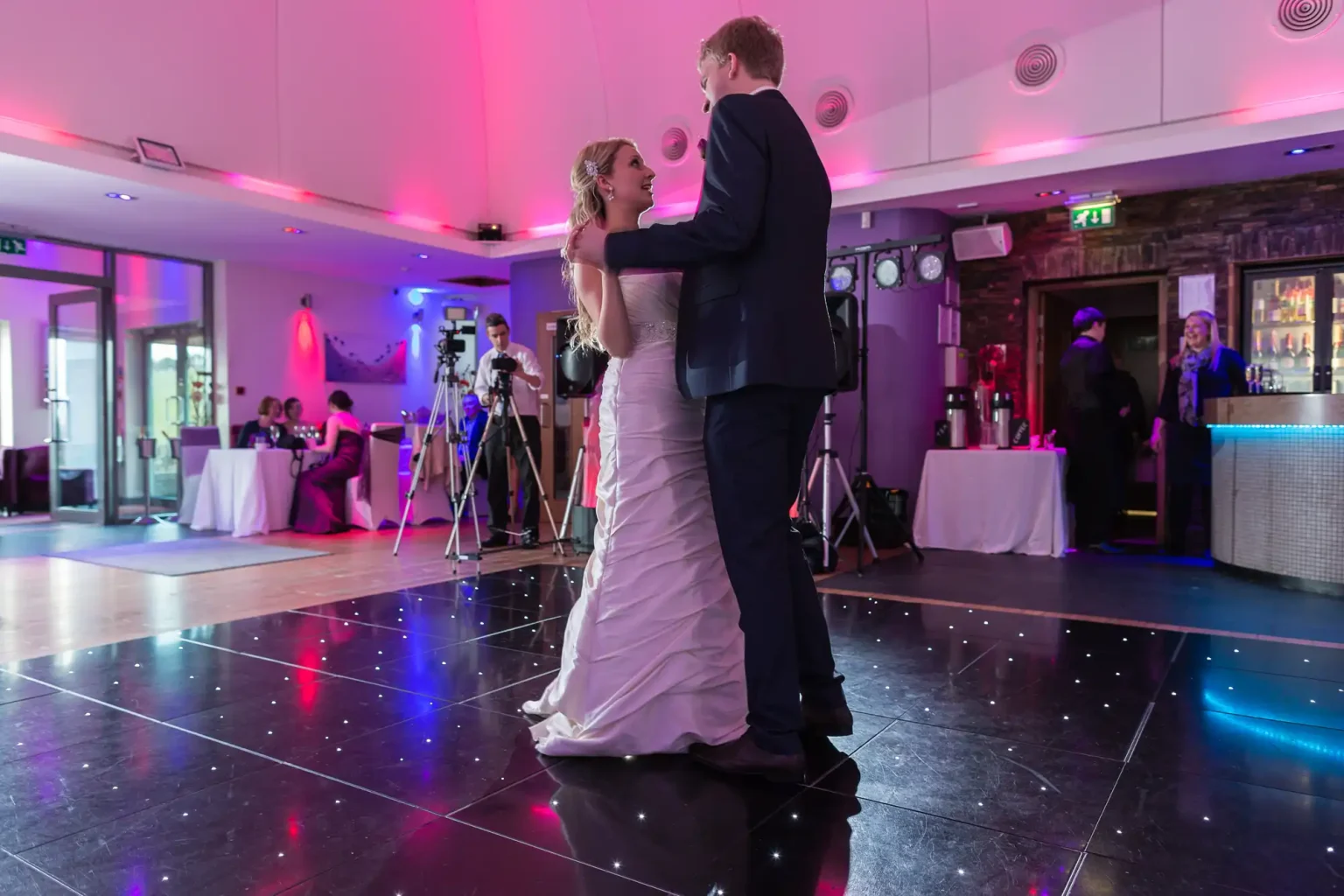 A bride and groom share a dance in a dimly lit venue with pink lighting, surrounded by guests.