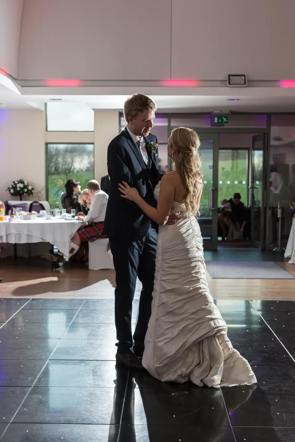 A bride and groom share a dance in a dimly lit banquet hall, with pink uplighting and guests in the background.