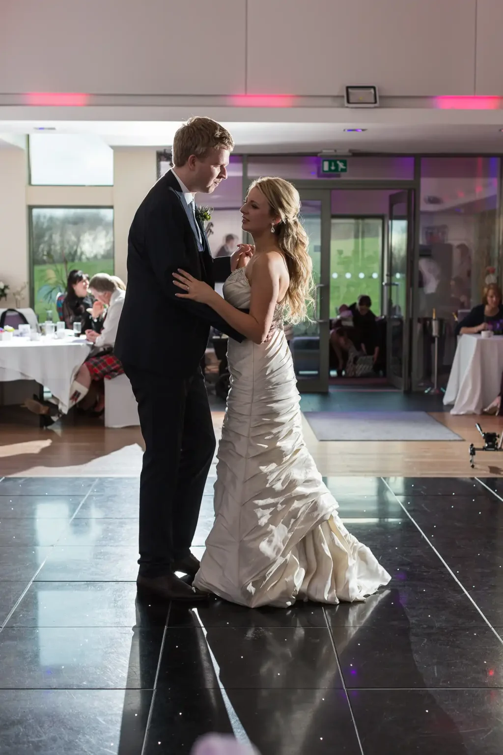 A bride and groom share a dance in a banquet hall with guests seated in the background, under pink lighting.