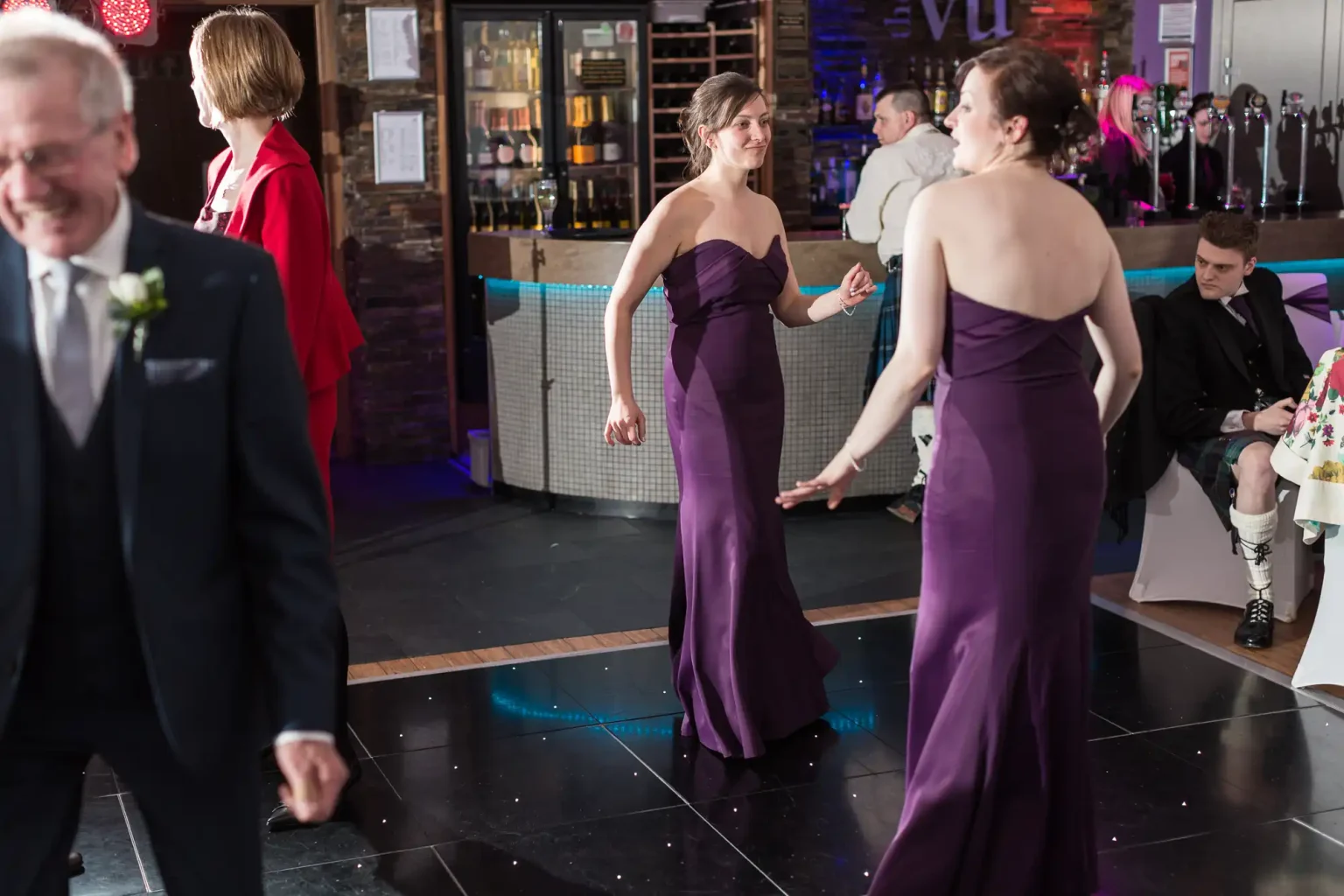 Two women in elegant purple dresses talking at a lively party with other guests in the background.