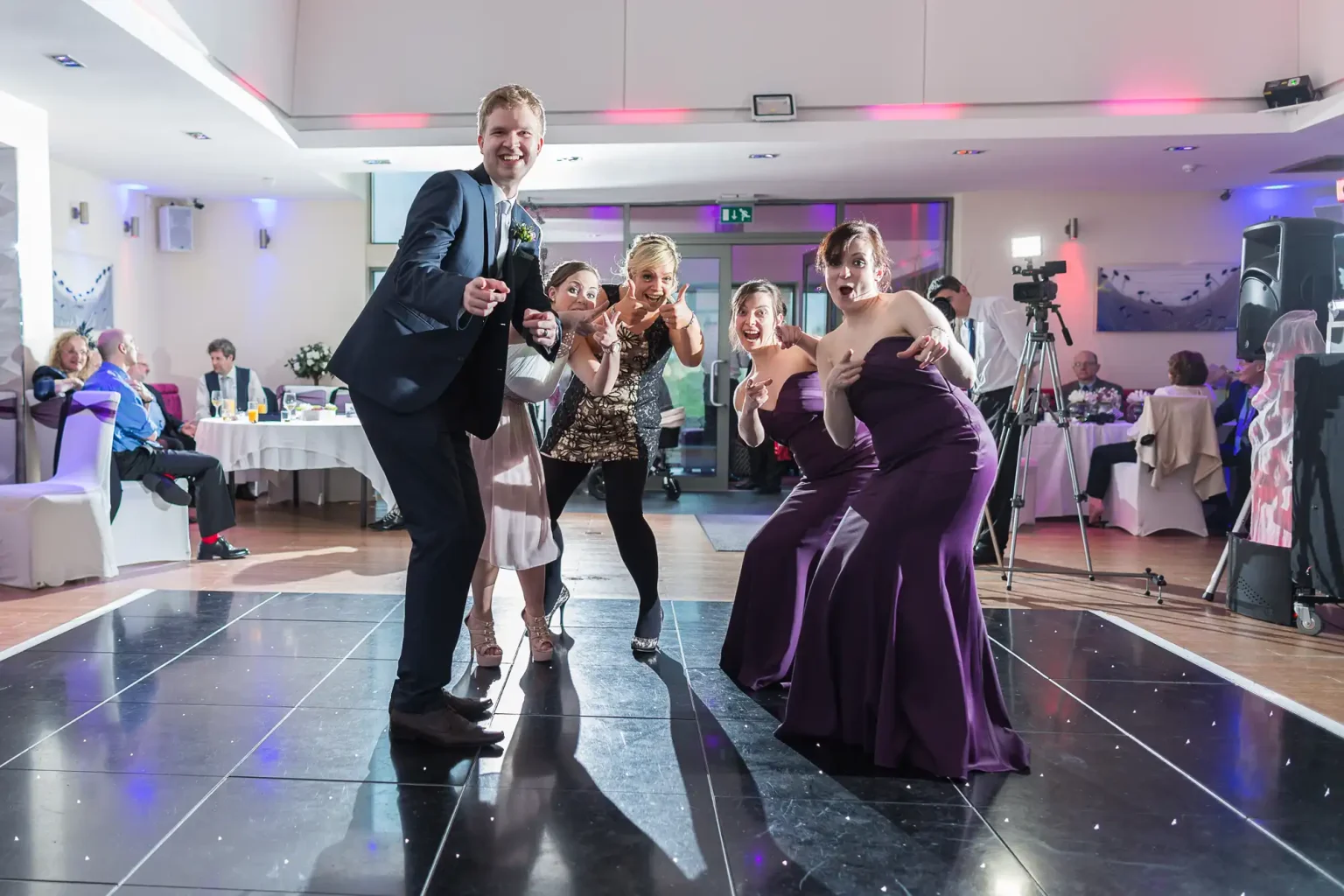 A group of people joyously dancing at a wedding reception, with one man playfully pointing at the camera.