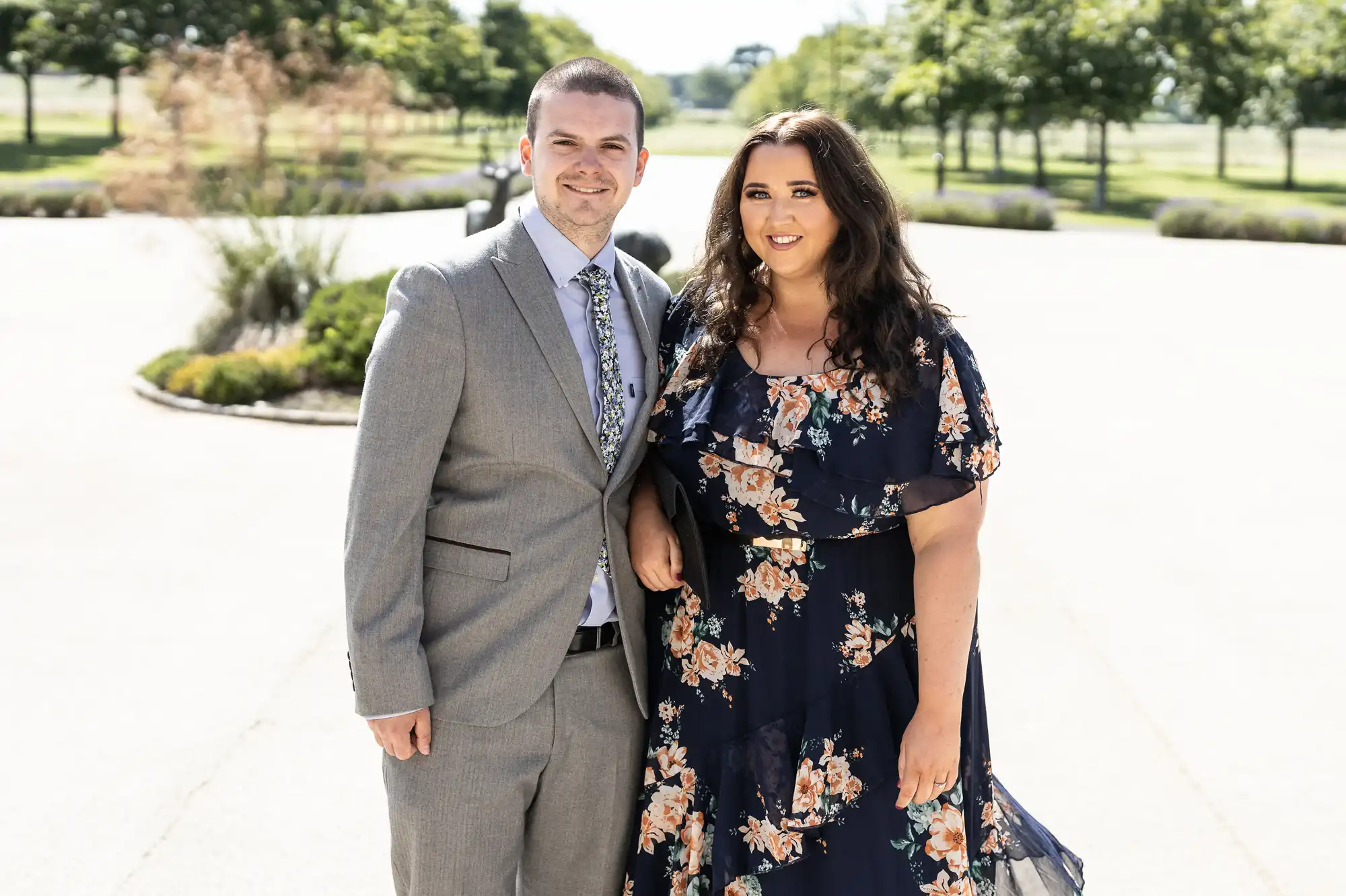 A man and a woman standing outdoors, smiling, dressed in formal attire with the man in a gray suit and the woman in a floral dress.