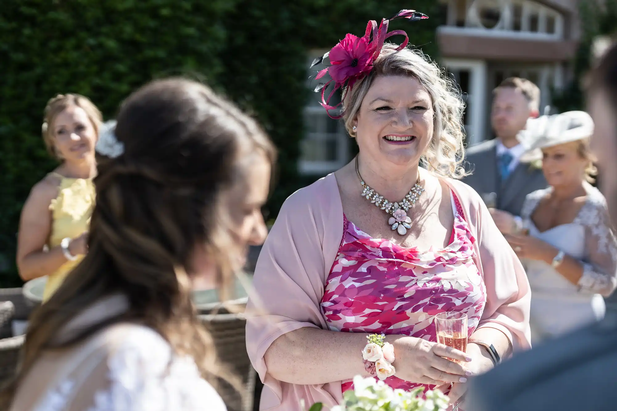 A woman in a pink floral dress and matching hat smiles while holding a champagne glass at a sunny outdoor wedding reception.