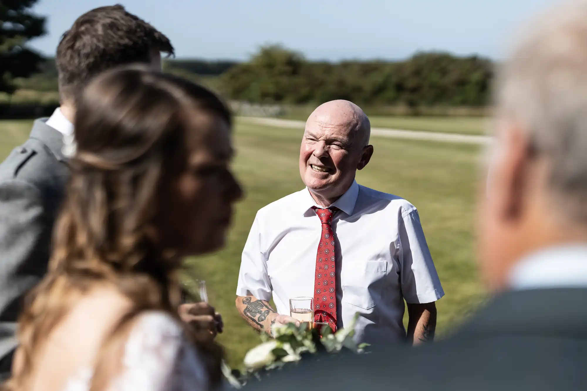 An older man in a white shirt and red tie laughing during a conversation at an outdoor wedding, with other guests around him.