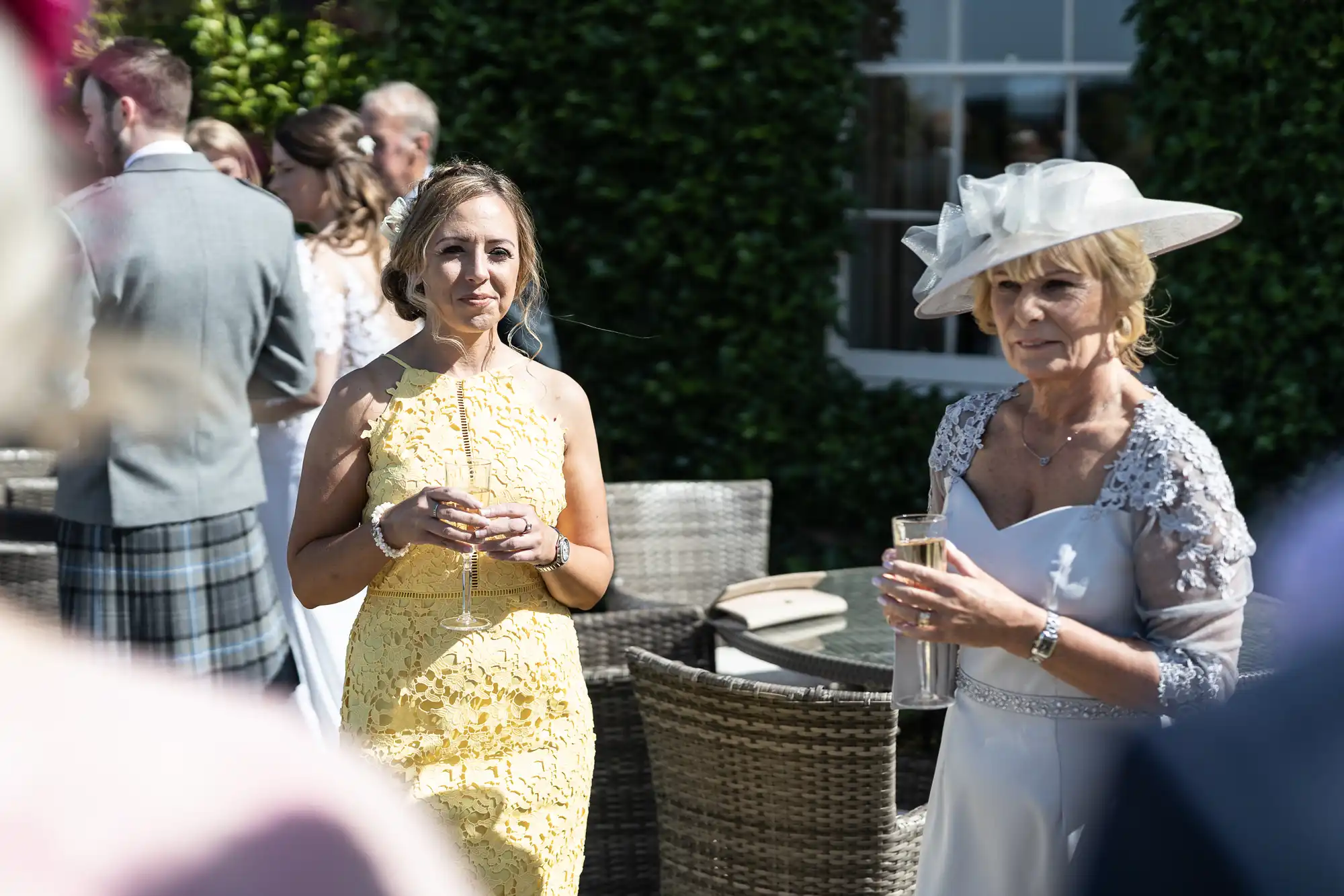 Two women at a social gathering outdoors, one in a yellow dress holding a drink, the other in a light blue outfit with a hat, also holding a drink.
