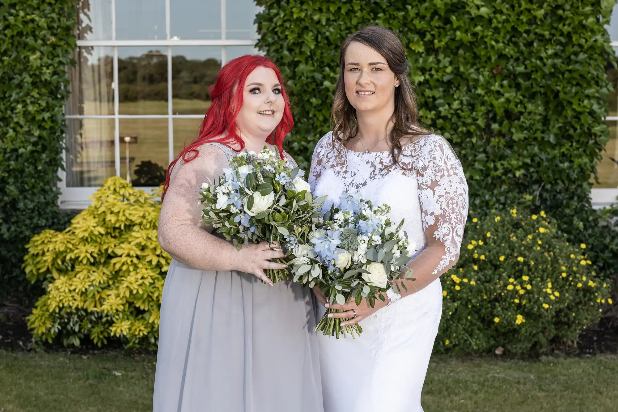 Two women in formal dresses holding bouquets stand together in front of a vine-covered building.