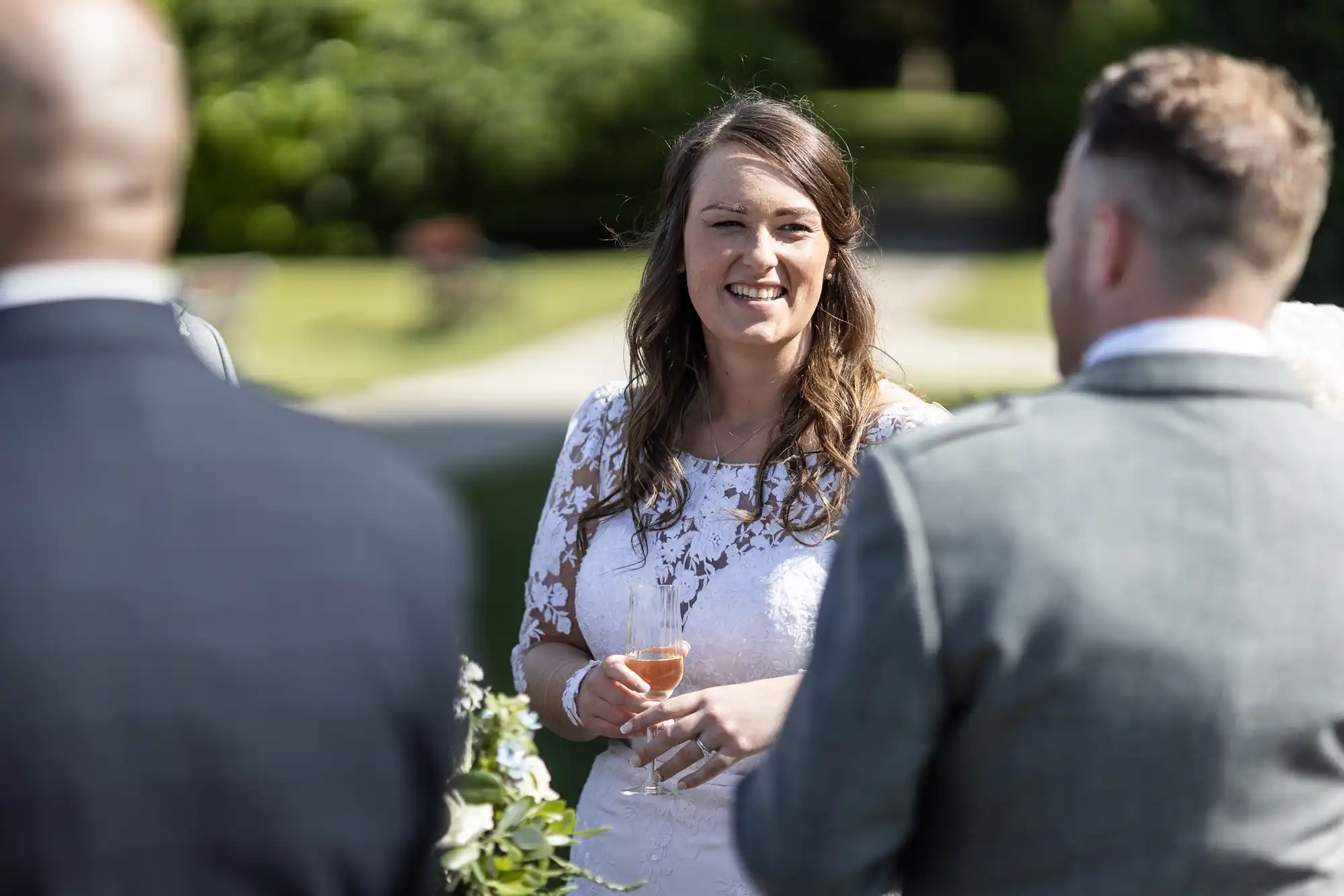A woman in a white lace dress holding a champagne flute smiles and talks to two men at an outdoor social gathering.