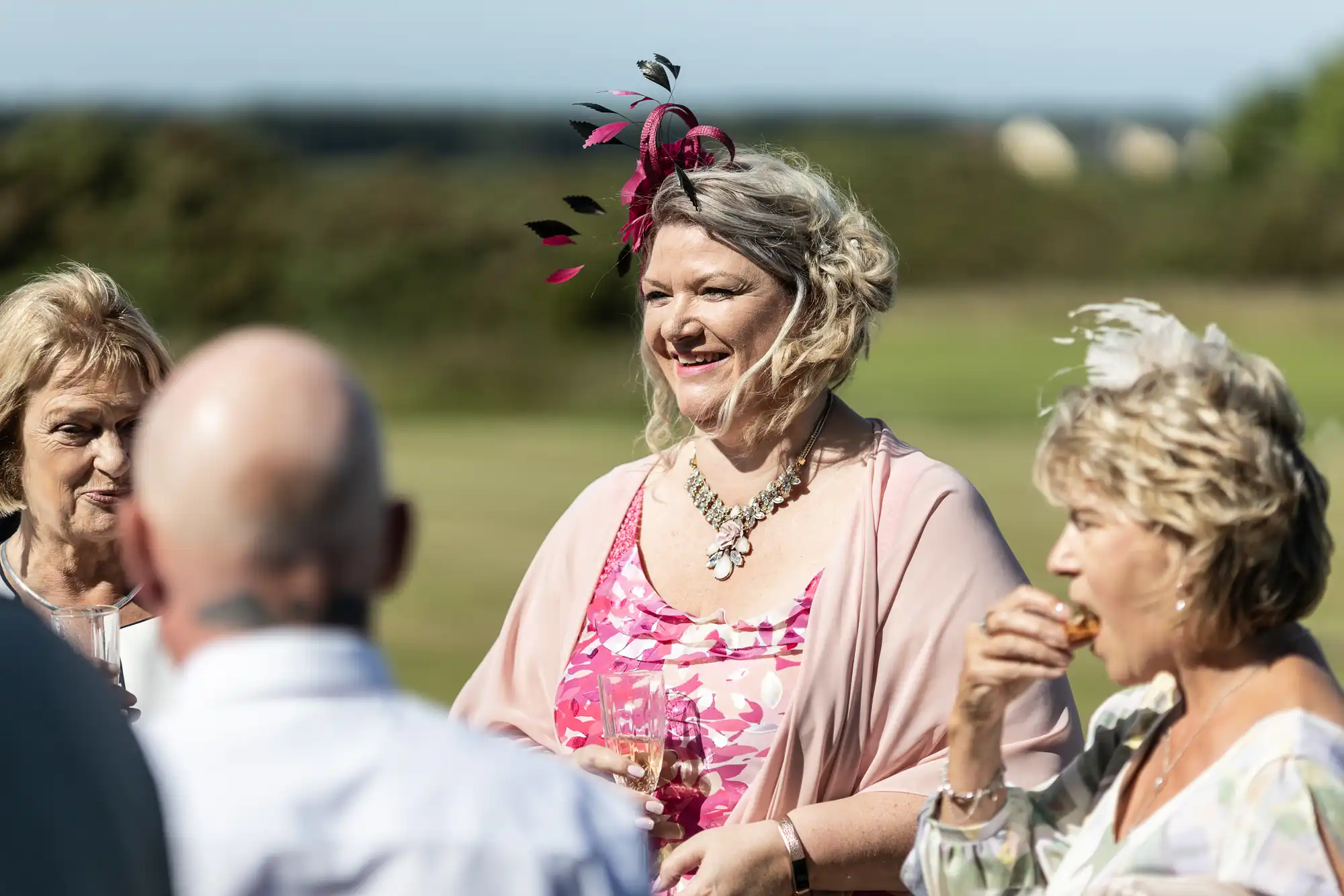 Three older women and one man socializing outdoors, one woman wearing a pink outfit with a feathered hair accessory, holding wine glasses.
