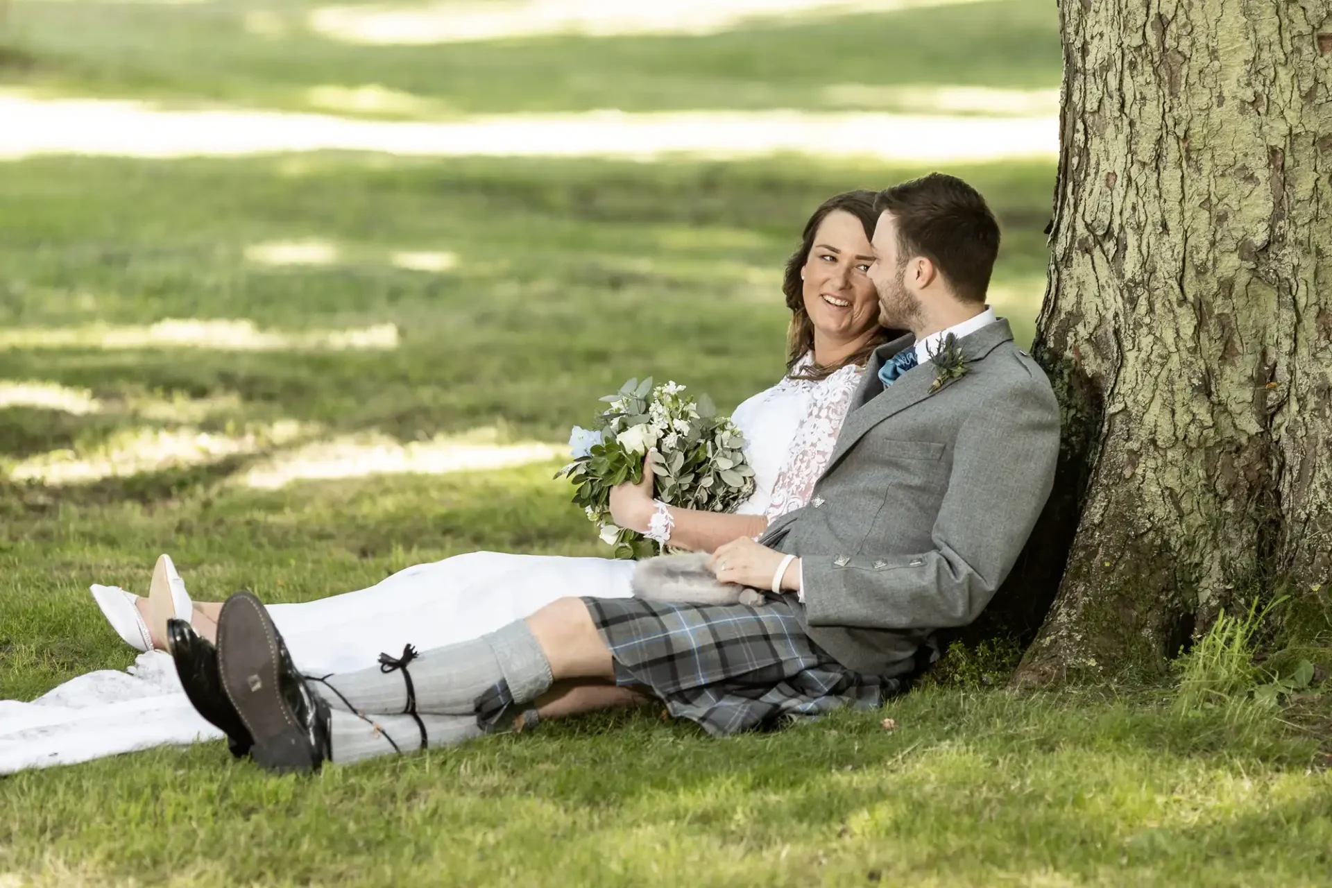 Archerfield House wedding photographer:
A newlywed couple in wedding attire smiling and sitting under a tree, with the groom wearing a kilt and the bride holding a bouquet.