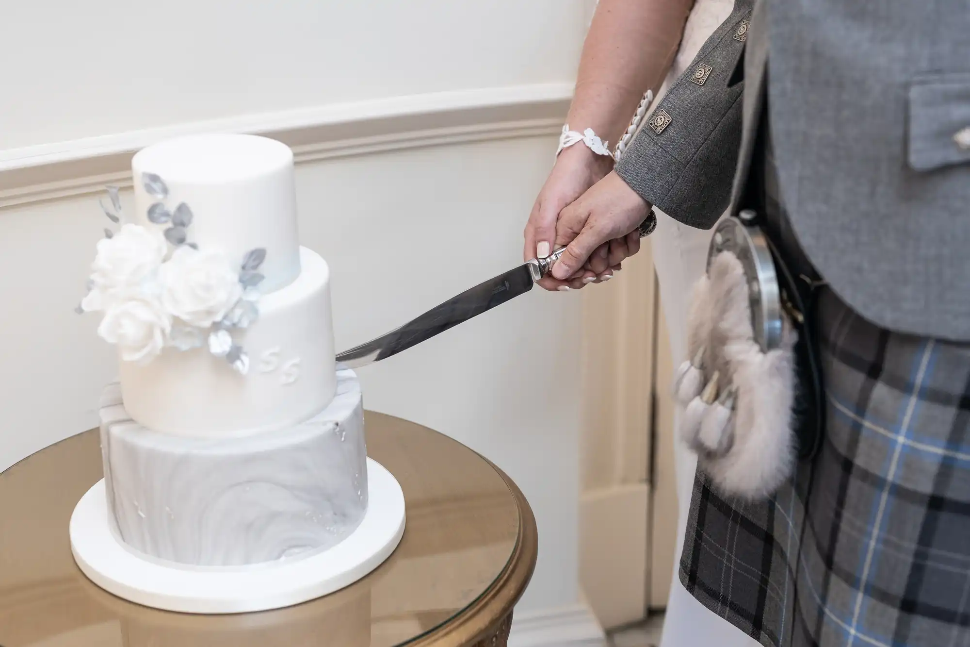 Two people in formal attire, one wearing a kilt, cutting a white floral-decorated cake with a large knife.