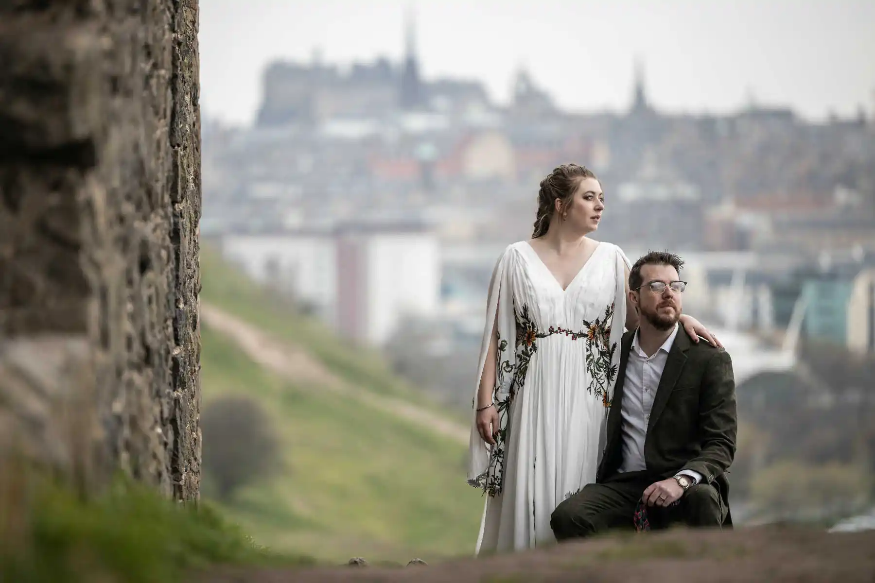 Arthur's Seat - Ginni and Henry: A woman in a white dress stands beside a man in a dark suit kneeling, with a blurred cityscape in the background.