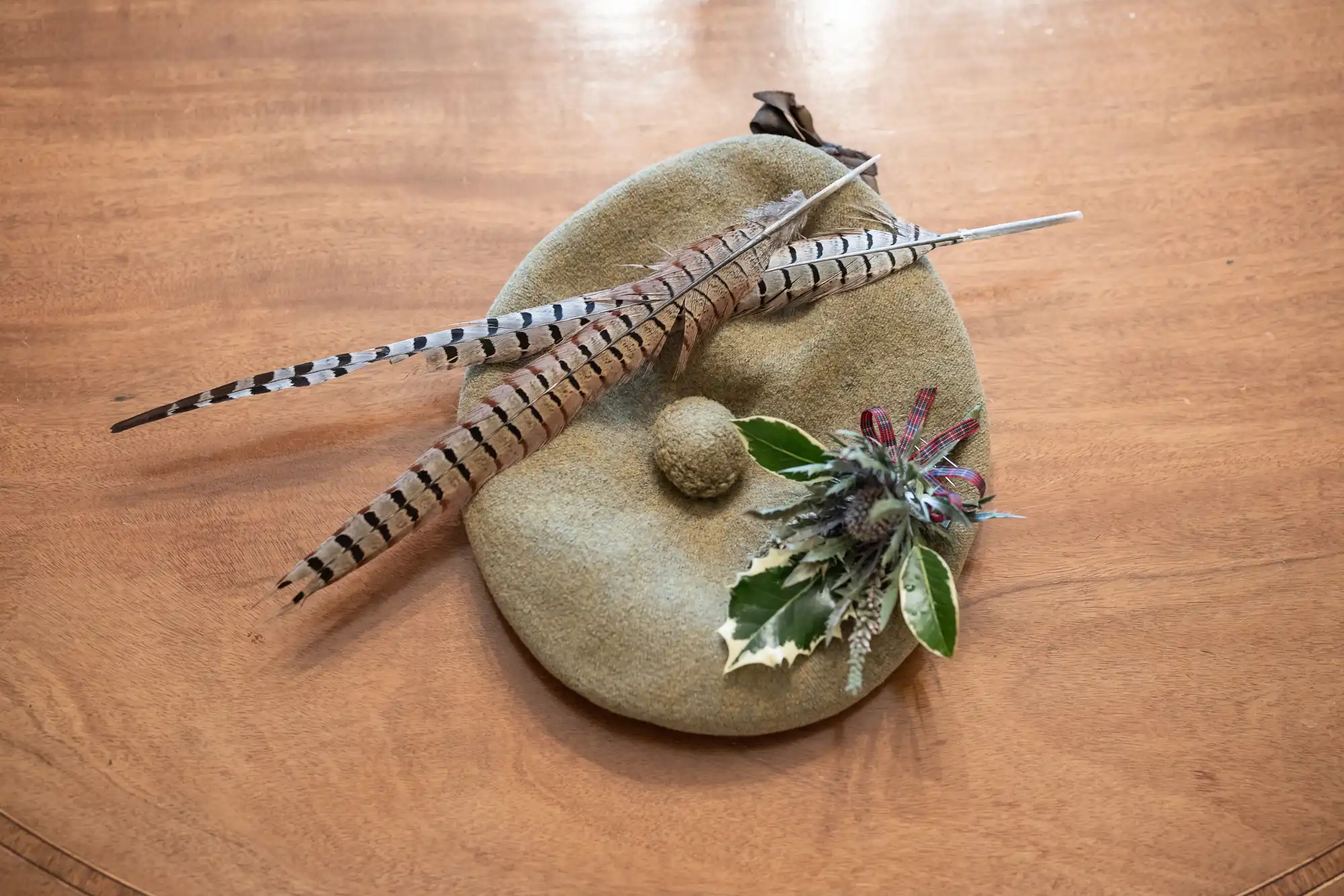 A beige beret adorned with two striped feathers and a small arrangement of greenery and flowers is placed on a wooden surface.