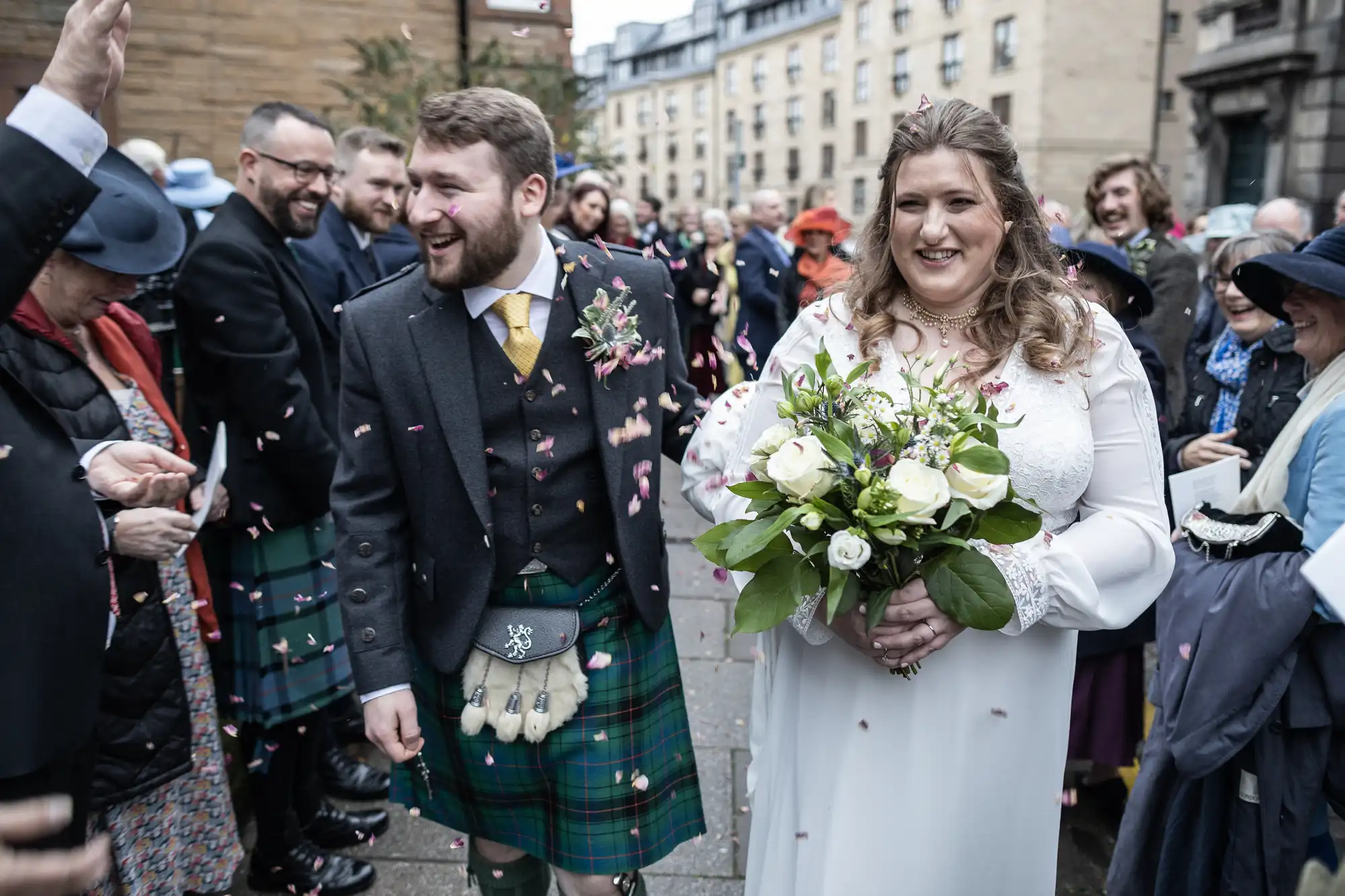 A bride and groom walk through an outdoor crowd, dressed in traditional Scottish attire. The groom wears a kilt, and the bride holds a bouquet. Guests throw confetti in celebration.