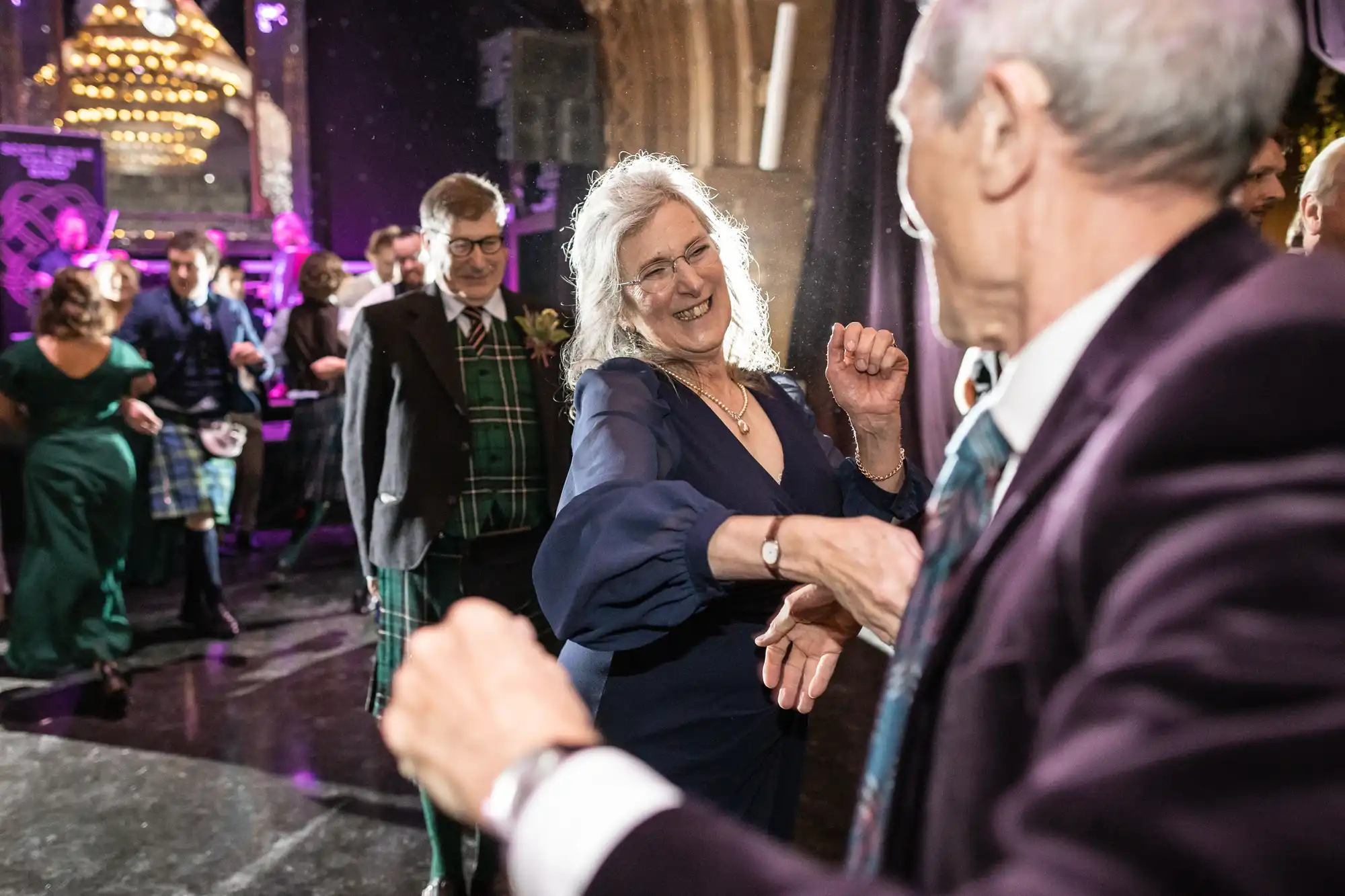 Ghillie Dhu wedding reception. A group of people dressed in formal attire are dancing and socializing at an indoor event with colorful lights in the background.
