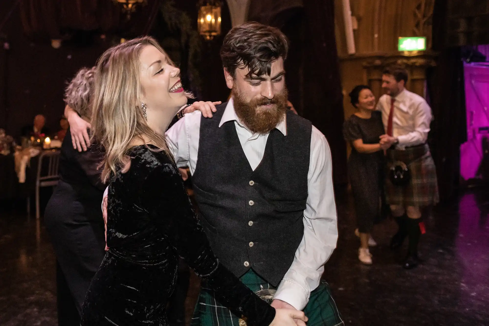 Ghillie Dhu wedding reception image of a woman and man, both smiling and holding hands, dance together in a dimly lit room. Other people are dancing in the background.