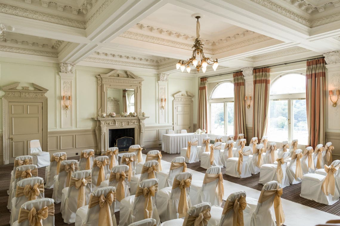 Esk Suite: Elegant wedding ceremony setup in a vintage-style room with rows of white chairs adorned with gold sashes, facing an ornate fireplace and a chandelier hanging from the ceiling.