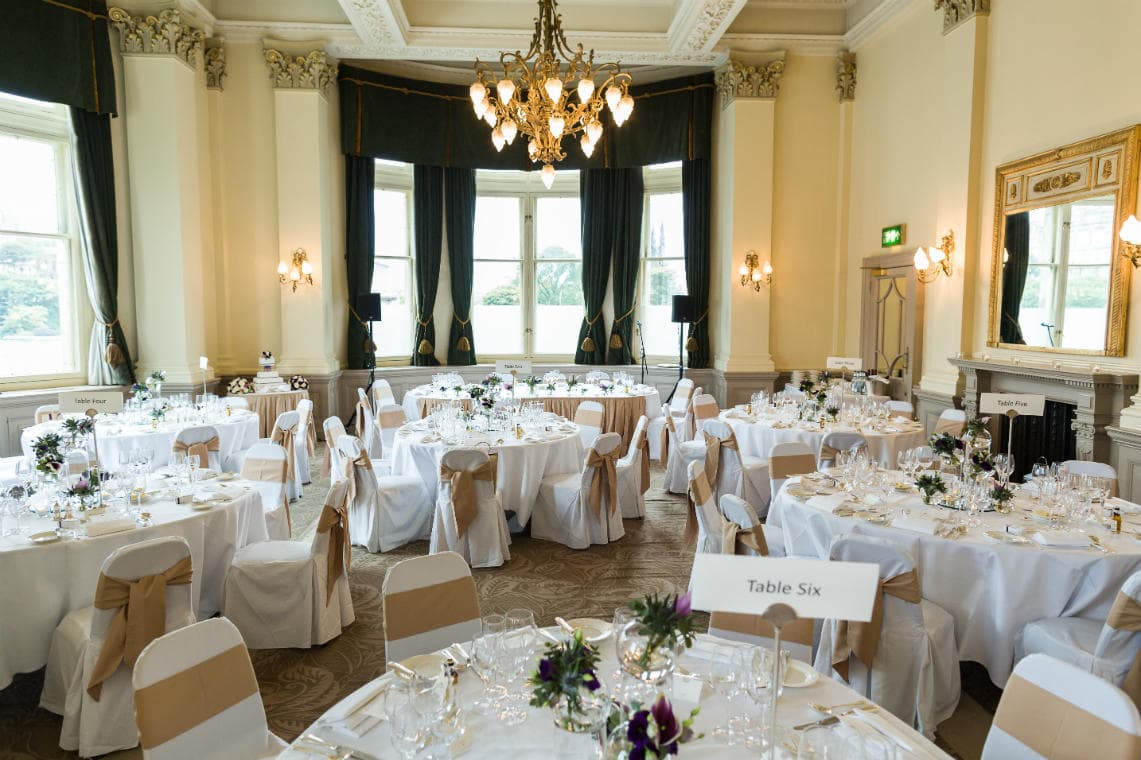 Holyrood Suite - set up for a wedding meal: a ballroom set up for an event with round tables covered in white cloths, decorated with floral centerpieces and place settings. Chandelier lights hang from the ceiling. Table numbers are visible.